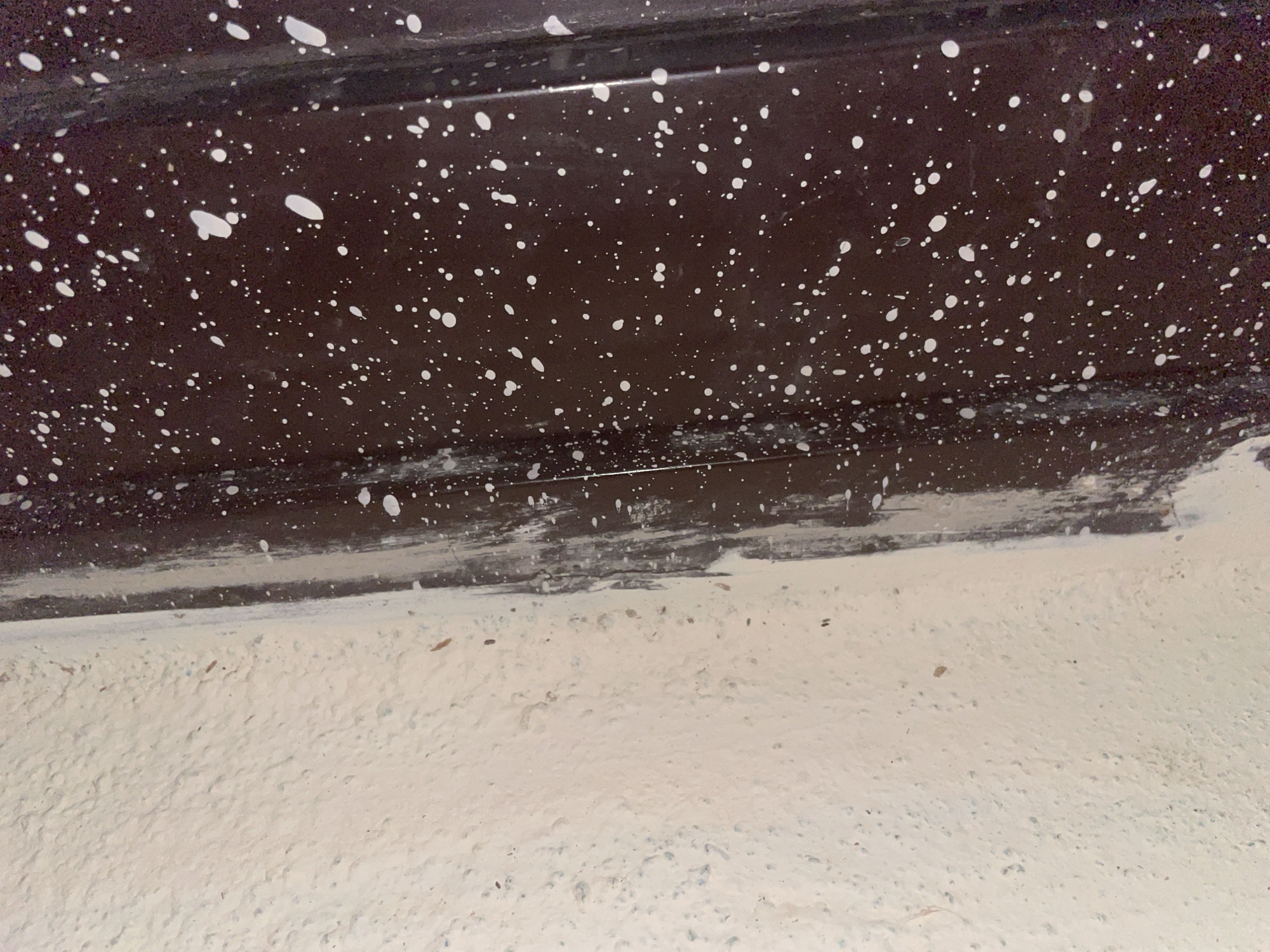 The image shows a close-up view of a textured, light-colored wall on the left and a dark, speckled surface on the right. The speckled surface appears to be a painted door frame or a similar architectural element, with numerous small white paint splatters against a dark brown background. There is a vertical seam or edge where the wall meets the speckled surface, with some white paint dripping or smudged along this transition. The lighting suggests an indoor environment. The location in the city of Jalingo, Nigeria, cannot be determined from the visual information provided in the image.
