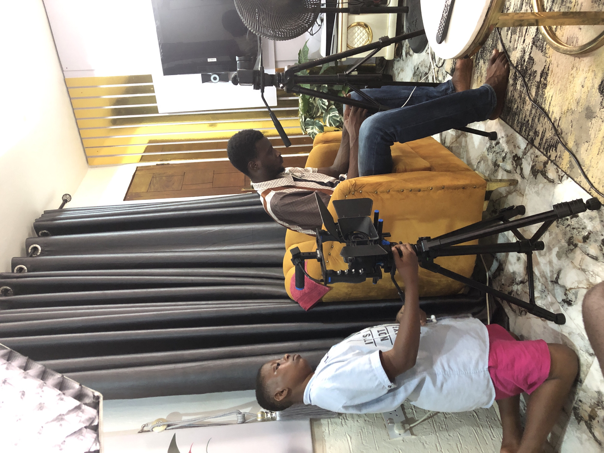 The image captures an indoor setting, likely a home or small studio, where a video recording or interview is taking place. The scene is dominated by two individuals and various filming equipment.
In the mid-ground, a man with short dark hair and a beard is seated comfortably in a mustard yellow armchair. He wears a patterned white and brown short-sleeved shirt and blue jeans, with his bare feet resting on a patterned rug. He is looking towards the left of the frame, seemingly at the camera operator.
In the foreground, a younger person (child or teenager) is positioned on the floor, operating a camera rig mounted on a small tripod. They are wearing a white t-shirt with partially visible text that reads "IT'S NOT A" and pink shorts. Their focus is directed upwards, towards the seated man, indicating they are actively filming him.
The room features a decorative background: a wall adorned with horizontal gold-colored panels, and a large television screen mounted above them. Dark grey curtains are visible on the left side of the frame. To the right of the seated man, a white round side table holds a remote control, and a black stand-up fan is partially visible behind it. Another tripod or light stand is positioned behind the armchair. The floor is covered with a light-colored patterned rug, and a power strip is visible near the person on the floor. The overall setup suggests a deliberate effort for a professional or semi-professional video production.