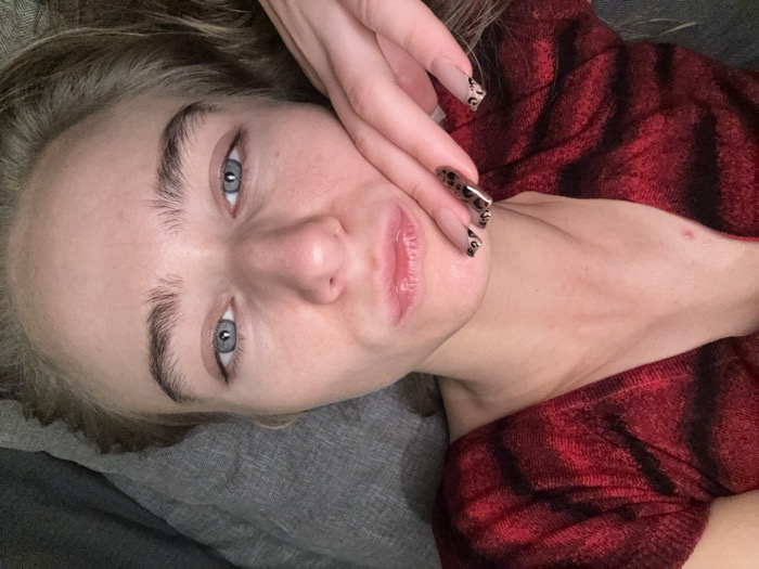A woman with fair skin, light blonde hair, and striking blue eyes gazes into the camera. She wears a red and black plaid shirt. Her hand, adorned with leopard-print nails, rests on her cheek. The background is a soft, neutral gray, suggesting a couch or bed. Her expression is serene, lips slightly parted.