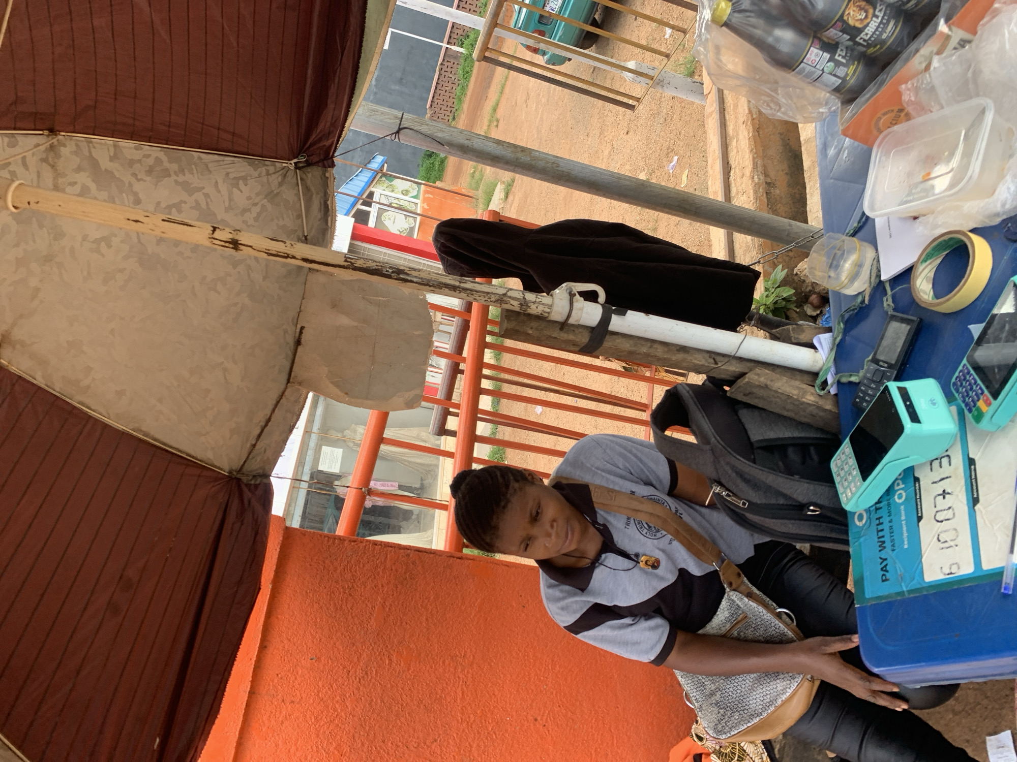 This image depicts an outdoor street or roadside stall in Jos, Nigeria, during daylight hours. A young woman, likely a vendor, is seated behind a blue table, positioned under a temporary canopy made of brown and grey fabric. She wears a grey and black polo shirt with text including "TRIN," "TRIA," and "HOLY TR" visible, along with dark pants and a light-colored shoulder bag. Her gaze is directed downwards, and her hand is near the table's surface.
The blue table is densely covered with various items indicating a small business operation. These include several bottles of "Fearless" energy drink in a plastic bag, clear plastic containers, a roll of masking tape, a blue pen, and two mobile phones (one a classic flip phone, the other a more modern type). Prominently displayed are two Point-of-Sale (POS) terminals, one of which is a distinctive mint green color. A document or sticker on the table features text such as "PAY WITH TH," "FASTER & MORE," "Recipient Bank OP," and a number "6100713.," followed by "Account Name," suggesting financial transaction services are offered. A grey backpack is also visible behind the table items.
The background reveals a dusty ground, an orange textured wall, and a metal railing. Beyond these, glimpses of other buildings and a light green car parked in the distance reinforce the outdoor, semi-urban setting. The bright lighting indicates it is daytime.