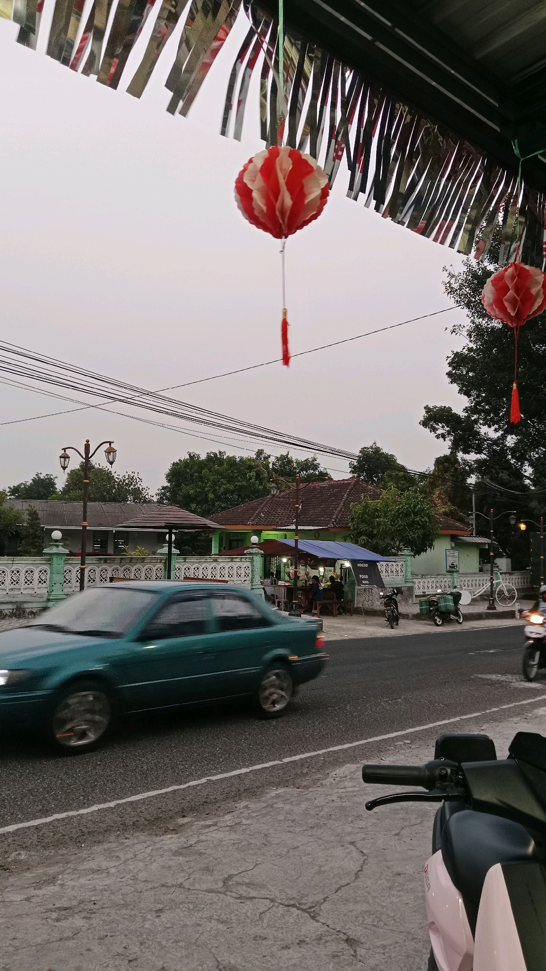 The image captures a street scene in Baron, Indonesia, with a focus on a roadside food stall. In the foreground, a teal-colored car is captured in motion, blurring as it drives past. The pavement in front of the stall shows cracks. Behind the car, a street lamp with two lights stands beside a green fence with white decorative elements. The food stall itself is covered by a blue awning and has tables and benches where several people are seated. A sign above the stall reads "NDESO FOOD & DRINK." To the right of the stall, two motorcycles are parked, one green and one black. A white bicycle is also parked nearby. Further to the right, another motorcycle is in motion. Overhead, red and white paper lanterns are hanging, along with silver tinsel. The sky is overcast, suggesting either dawn, dusk, or a heavily clouded day. In the background, trees and houses are visible.