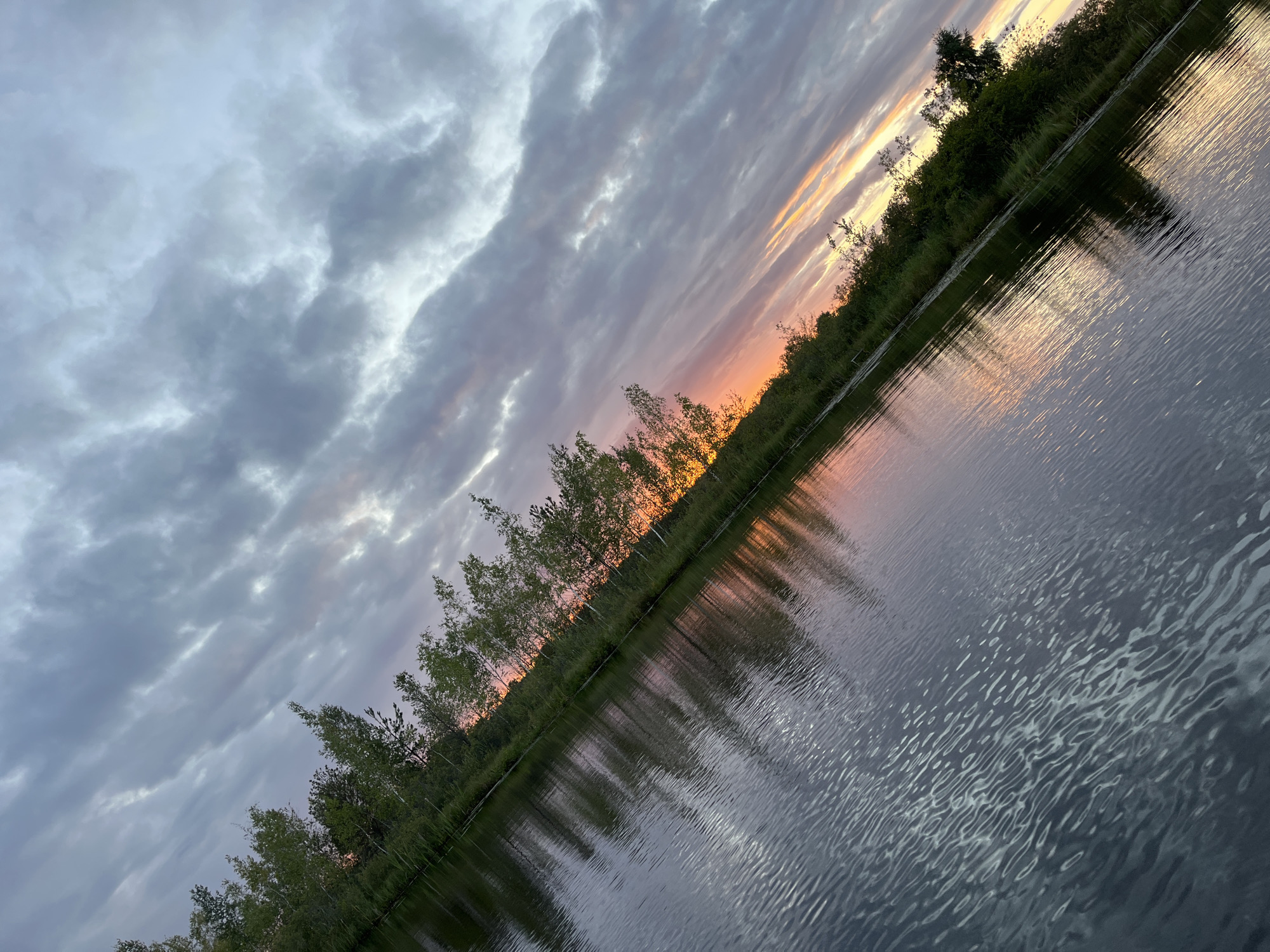 This image shows a body of water with a tree-lined shore under a cloudy sky. The water reflects the sky and the trees. The sun appears to be setting, casting an orange and pink glow on the horizon, visible between the trees. The scene is tilted, giving it a dynamic perspective. The location is identified as Dunilavichy Village Council, Belarus.