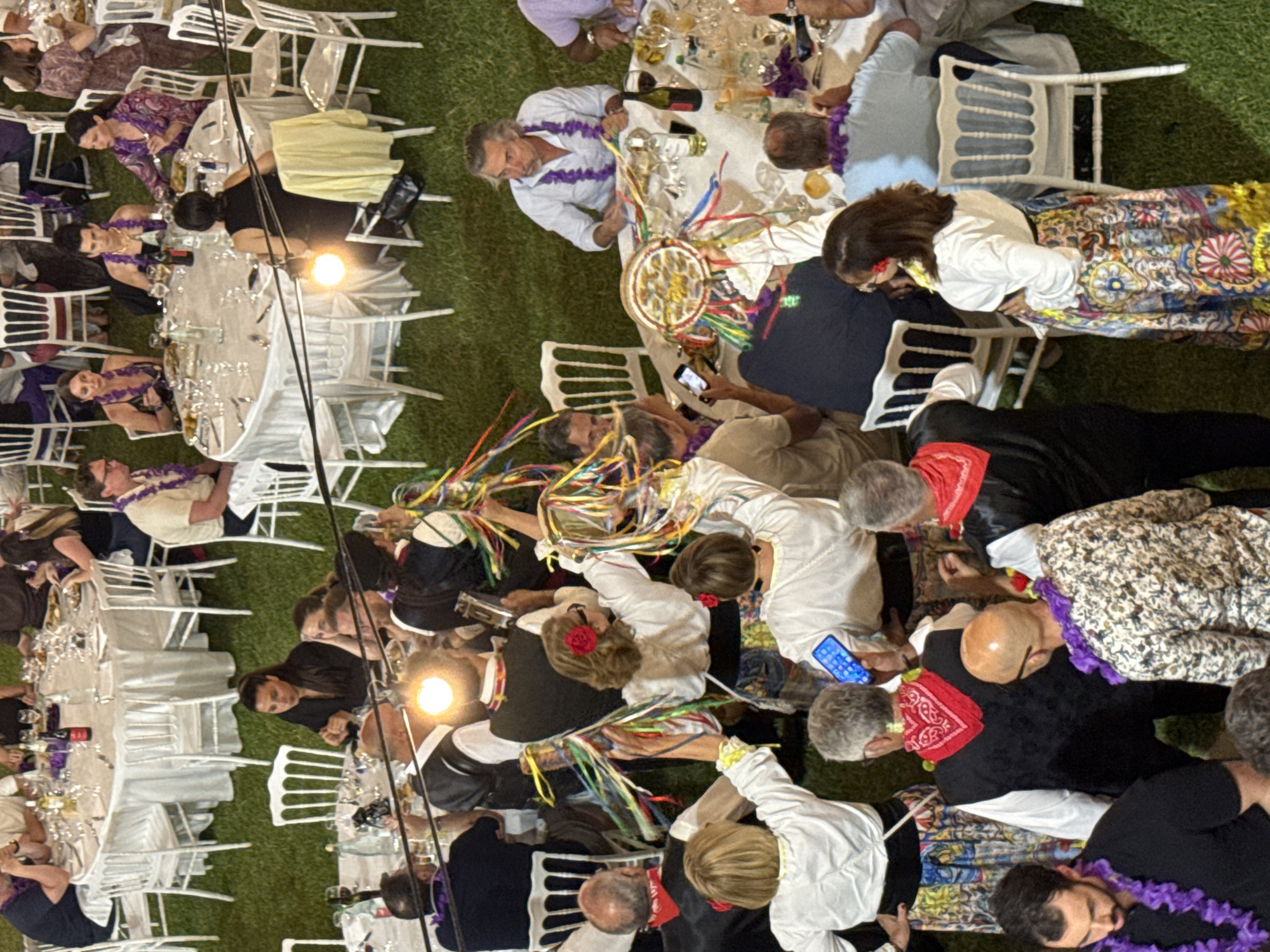 This forensic description details an outdoor evening event occurring on a green grass lawn in Capaci, Italy. Numerous adults are gathered around multiple round tables covered with white tablecloths and set with white chairs. Each table features place settings, wine glasses, bottles of wine (red and white), and other beverages.
In the upper half of the image, seated guests are observed dining and socializing. Many individuals wear purple or white leis around their necks. Specific individuals include:
* At a central table in the upper half, a woman in a black top is seated with her back to the viewer, facing three other women across the table. Two of these women wear purple leis with their attire, one in a patterned dress and the other in a black top.
* To the right, a man with gray hair and a white shirt, also wearing a purple lei, is seated at a table, looking to his left.
The foreground and mid-ground are dominated by a group of individuals standing and participating in a festive activity. This group includes both men and women.
* Women wear white long-sleeved blouses, long multi-colored patterned skirts, and some have red flowers in their hair. They are actively holding and waving numerous long, colorful ribbons.
* Men wear white shirts, dark vests, and red bandanas or scarves around their necks. Some are also holding ribbons.
* One man in the lower left foreground is wearing a black shirt and a purple lei. Another man in the lower right foreground wears a patterned shirt and a purple lei.
* A bright light source, possibly a flash or lamp, is visible in the center-left mid-ground, illuminating a man from behind who is holding a metallic object resembling a musical instrument.
* On a table in the center-right, a large, circular, decorative object resembling a dreamcatcher, adorned with numerous colorful ribbons, is present.
* Several individuals throughout the scene are observed holding or looking at mobile phones.
The overall environment is an open-air setting with artificial illumination consistent with a nighttime event. The ground is uniformly covered in short, well-maintained green grass. Interactions include dining, socializing, and an apparent celebratory performance or dance involving ribbons.