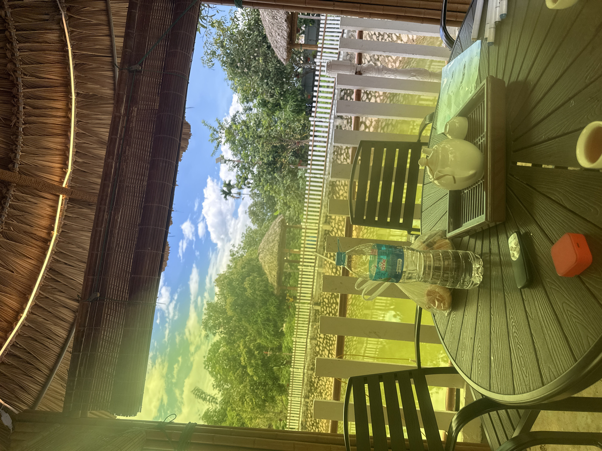 The image presents a detailed view from a semi-outdoor seating area, characteristic of a cafe or restaurant, situated in Nghia Lo Ward, Vietnam. No individuals are visible within the scene.
The foreground features a round, dark-toned table with a textured surface mimicking wood grain. Prominently displayed on the table is a clear plastic bottle of "LaVie" branded water, featuring a blue cap and a label indicating "1900 1906". Adjacent to the bottle, a clear plastic bag contains two light-colored baked goods. A black remote control with silver buttons and a small, square red object (possibly a portable speaker) rest on the table. A white ceramic teapot with a lid and a small matching cup are placed on a dark brown slatted wooden tray. To the right, a light blue folder or book and several white-bodied marker pens are visible. A ceramic cup holding a light brown liquid (consistent with tea) is partially cropped at the bottom center-right. Dark-colored chairs with slatted backs and armrests are partially visible around the table.
Overhead, a thatched roof constructed from dried organic material, likely straw or palm leaves, forms the ceiling. A continuous, thin illuminated strip, indicative of LED lighting, is installed along the interior edge of the thatch. A partially rolled-up woven bamboo or wooden blind hangs below the roof structure. Vertical structural supports, appearing to be bamboo poles, frame the viewing area on both the left and right.
Beyond the immediate seating area, a body of water with a distinct greenish-yellow reflection occupies the middle ground. A white picket fence, composed of vertical square posts and horizontal rails, borders the water. The background is dominated by lush green trees and dense foliage under a bright blue sky with scattered white cumulus clouds. Two distinct thatched-roof structures, resembling gazebos or small huts, are visible within the distant landscape, one on each side of the water body. A tall lamp post is discernible among the trees on the right side. Palm trees are identifiable within the vegetation on the left side of the scene.
