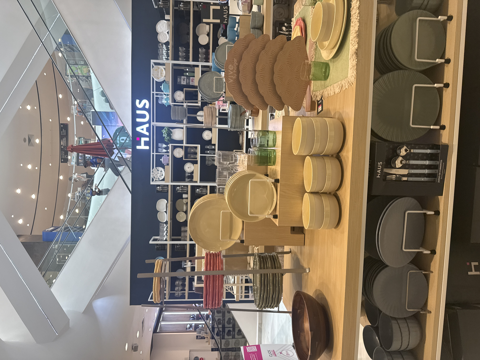 Here's a detailed description of the image:
**Setting:**
The image captures the interior of a HAUS store, likely a department store featuring homewares and kitchenware. Based on the signage and product offerings, this appears to be a home goods section within a larger department store. The presence of a modern architectural element with glass railings and escalators indicates it is likely located in a shopping mall. The store is located in Puebla, Mexico.
**Objects:**
* Display shelves and tables showcasing a variety of dinnerware sets, including plates, bowls, cutlery, and placemats.
* Stacks of plates in different colors (tan, green, grey) and materials are arranged neatly.
* A wooden bowl is placed on the lower shelf.
* Glassware items and ceramic bowls are also on display.
* A cutlery set is packaged in a black box.
* Promotional materials are visible, including signage reading "HAUS" and a small sign with "CLICK" and "2 hrs)."
* The backdrop features shelves filled with additional tableware items.
**Environment:**
The store is brightly lit with modern fixtures. The background showcases an open architectural design with multiple levels and escalators.
**Overall Impression:**
The image depicts a well-organized and attractive display of home goods in a modern retail setting.