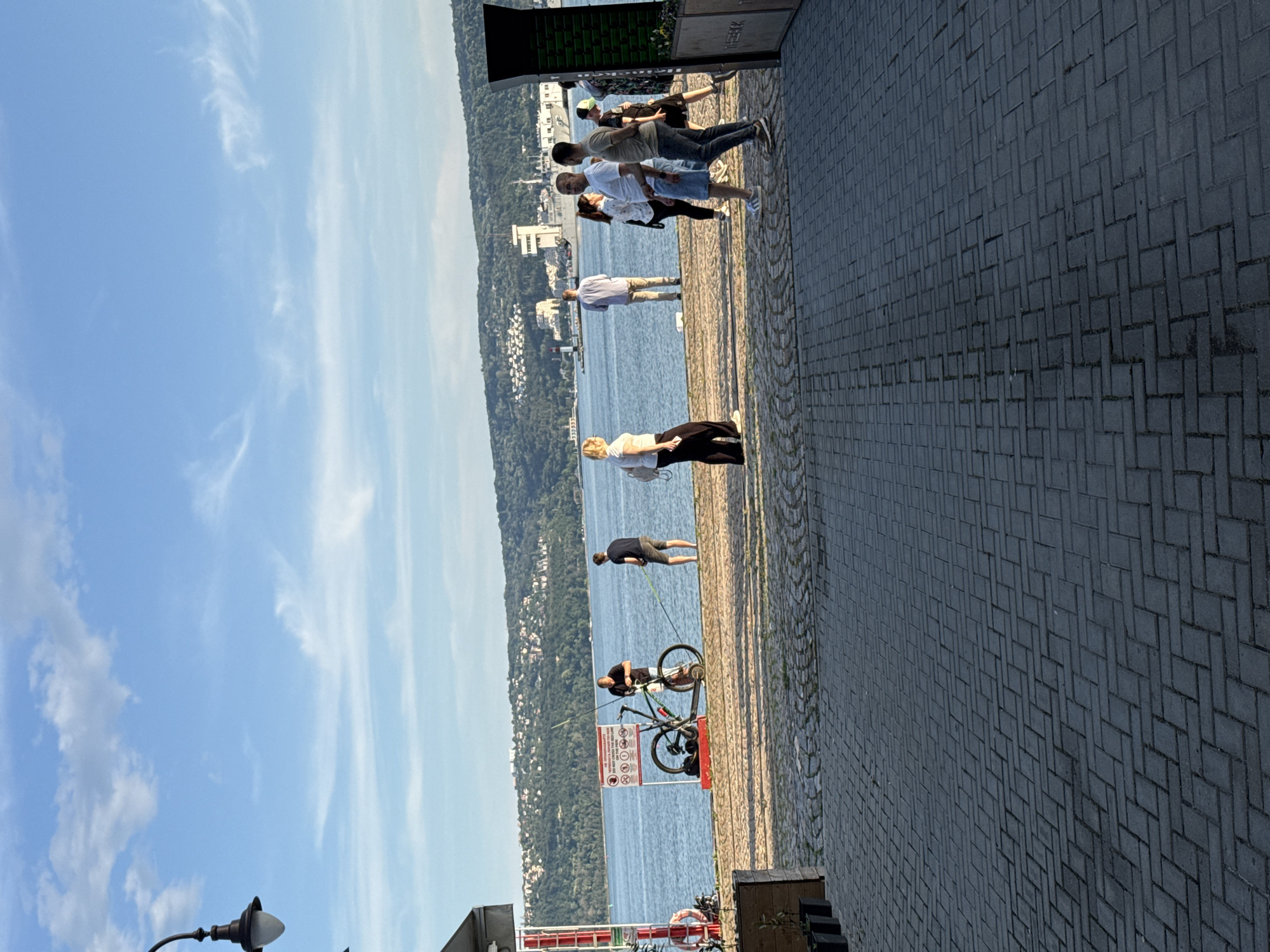 A group of people are gathered near a body of water on a paved walkway. A man is standing next to a bicycle and a fishing rod. Several other individuals are conversing or walking. In the background, there is a cityscape with buildings and a hillside covered in trees. The sky is blue with some clouds.