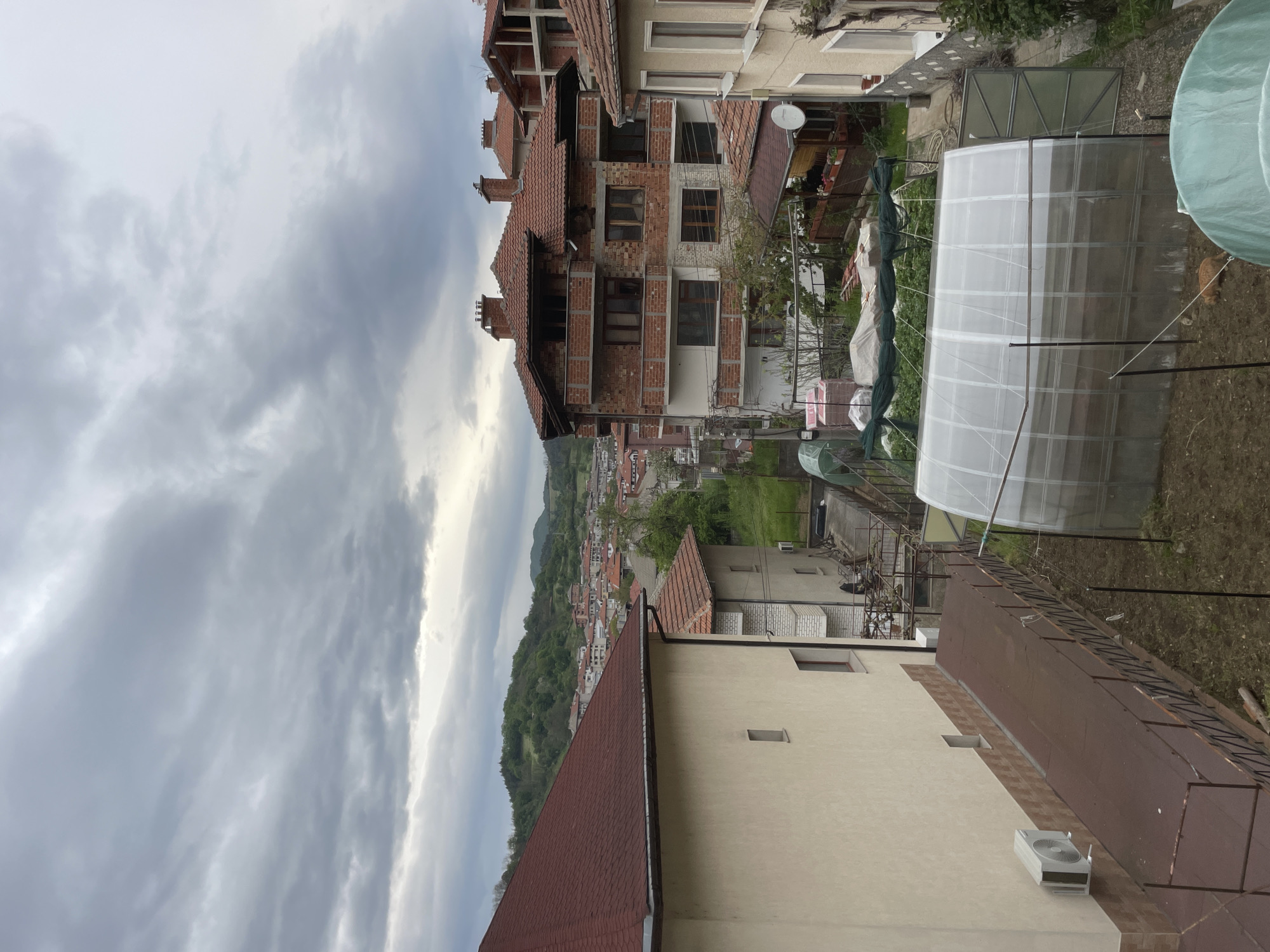 The image captures an outdoor scene in Zlatograd, Bulgaria, under a cloudy sky. The foreground features a section of a building with a light-colored exterior and a red-tiled roof. An air conditioning unit is visible on the side of the building. A greenhouse structure is located nearby. The middle ground shows multiple buildings with red tile roofs, a partially built brick building and lush green trees and grass. In the background, a hilly landscape and a dense urban area are visible.