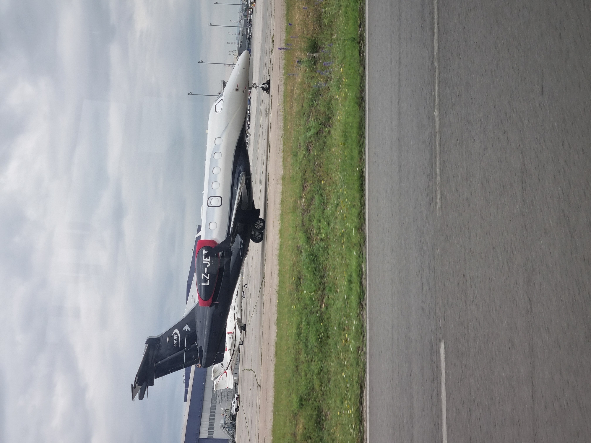 The image depicts a private jet, identified as LZ-JET, parked on an airport tarmac. The environment includes a grassy area adjacent to the tarmac, a runway marked with a white line, and an overcast sky. In the background, there are buildings and additional parked aircraft. Given the registration code LZ, the aircraft is likely located at Sofia Airport in Bulgaria.