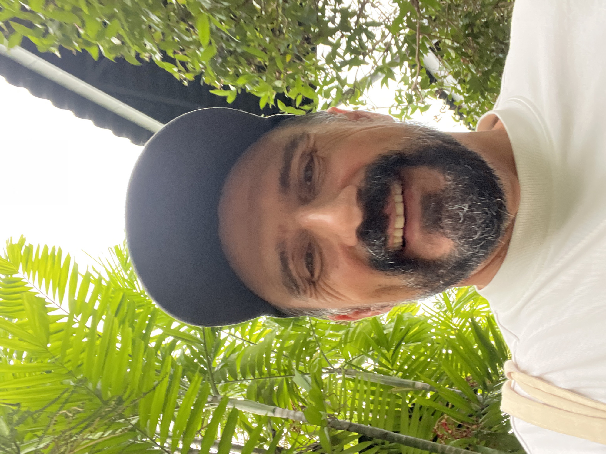 The photograph shows a man with a dark beard and a baseball cap, smiling broadly, with the camera angled upwards. He is wearing a white t-shirt. Behind him are green, leafy trees, and what appears to be the underside of a roof structure.
While the prompt mentions the city of บ้านบางรักษ์, Thailand, there is nothing in the visual content of the image to confirm that the photograph was taken there.