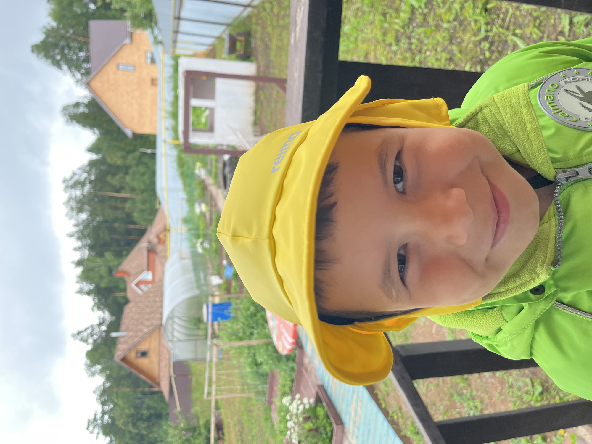 A close-up, outdoor, eye-level shot shows a young boy wearing a yellow hat and a bright green jacket. The boy is looking directly at the camera with a slight smile. The background is a slightly blurred view of a suburban or rural setting with houses, trees, and a greenhouse. The environment appears to be green and lush, suggesting a warm or temperate climate. The image does not provide any specific identifiable landmarks that would place it in the city of Chaikovsky, Russia.