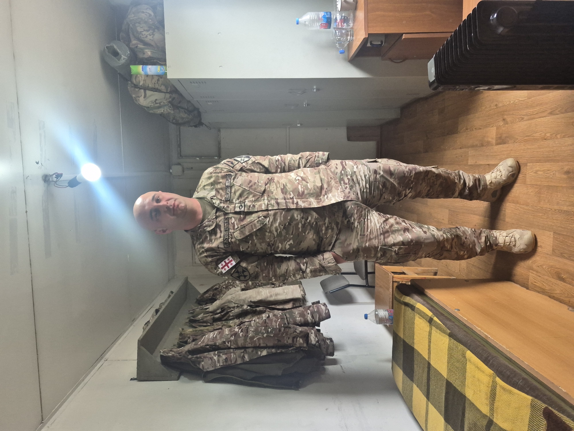 A man in military fatigues is lying in what appears to be a simple room, possibly a barracks, in Tbilisi, Georgia. He is wearing a camouflage uniform with a Georgian flag patch and name tag reading "GHUMASH." The room contains a wooden floor, a bed with a yellow and brown blanket, a black radiator, a water bottle, and some stacked clothing. The room is lit by a single light bulb.