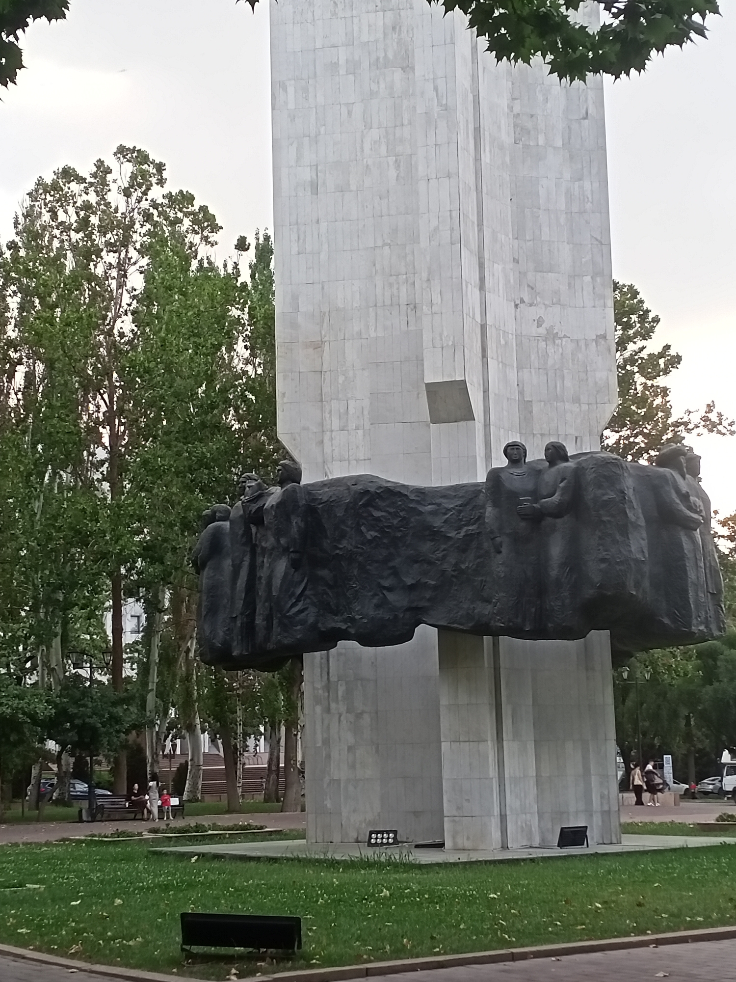 The image shows a monument with a large, dark, sculpted frieze featuring multiple figures. This frieze is positioned above a tall, light-colored, rectangular structure composed of many large, square tiles. The monument is situated in a park-like setting with a well-maintained lawn in the foreground. Several trees with lush green foliage surround the monument. In the background, there are more trees, buildings, and some vehicles, suggesting an urban environment. A few people are visible in the background, walking and sitting on benches. There is also some greenery and landscaping around the base of the monument.