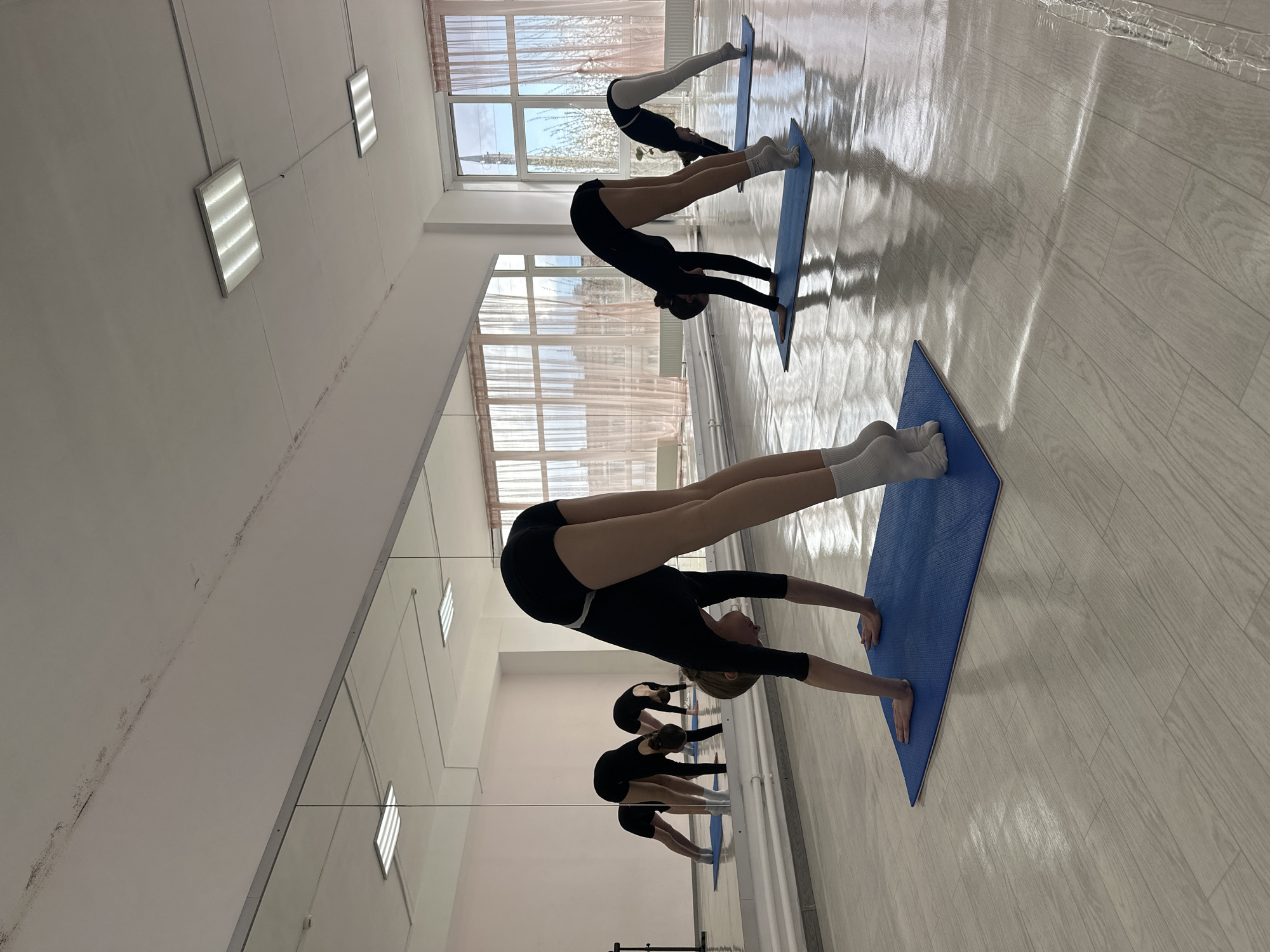 The image depicts an indoor gymnastics or yoga class taking place in a studio in Votkinsk, Russia, during daytime. The scene primarily features three young individuals, likely female, dressed in black leotards and white socks, performing a downward-facing dog or similar inverted V-shape stretch on blue yoga mats.
In the foreground, a participant is positioned with her hands flat on a mat and her head lowered, back arched, and legs extended upwards. Behind her, two other individuals are visible in similar poses, further into the room. A large wall-mounted mirror on the left reflects the participants and the studio, creating an illusion of more people and adding depth to the space.
The studio has a light-colored wooden floor that reflects the bright natural light pouring in from large windows on the far wall. These windows are partially covered by light, sheer curtains, revealing a clear sky and some distant scenery outside. The ceiling is white, fitted with several rectangular fluorescent light fixtures. Some minor wear and discoloration are noticeable on the upper walls and ceiling. The overall atmosphere suggests a focused and disciplined stretching or practice session, with the strong natural light highlighting the forms and movements of the participants. No discernible text is visible in the image.