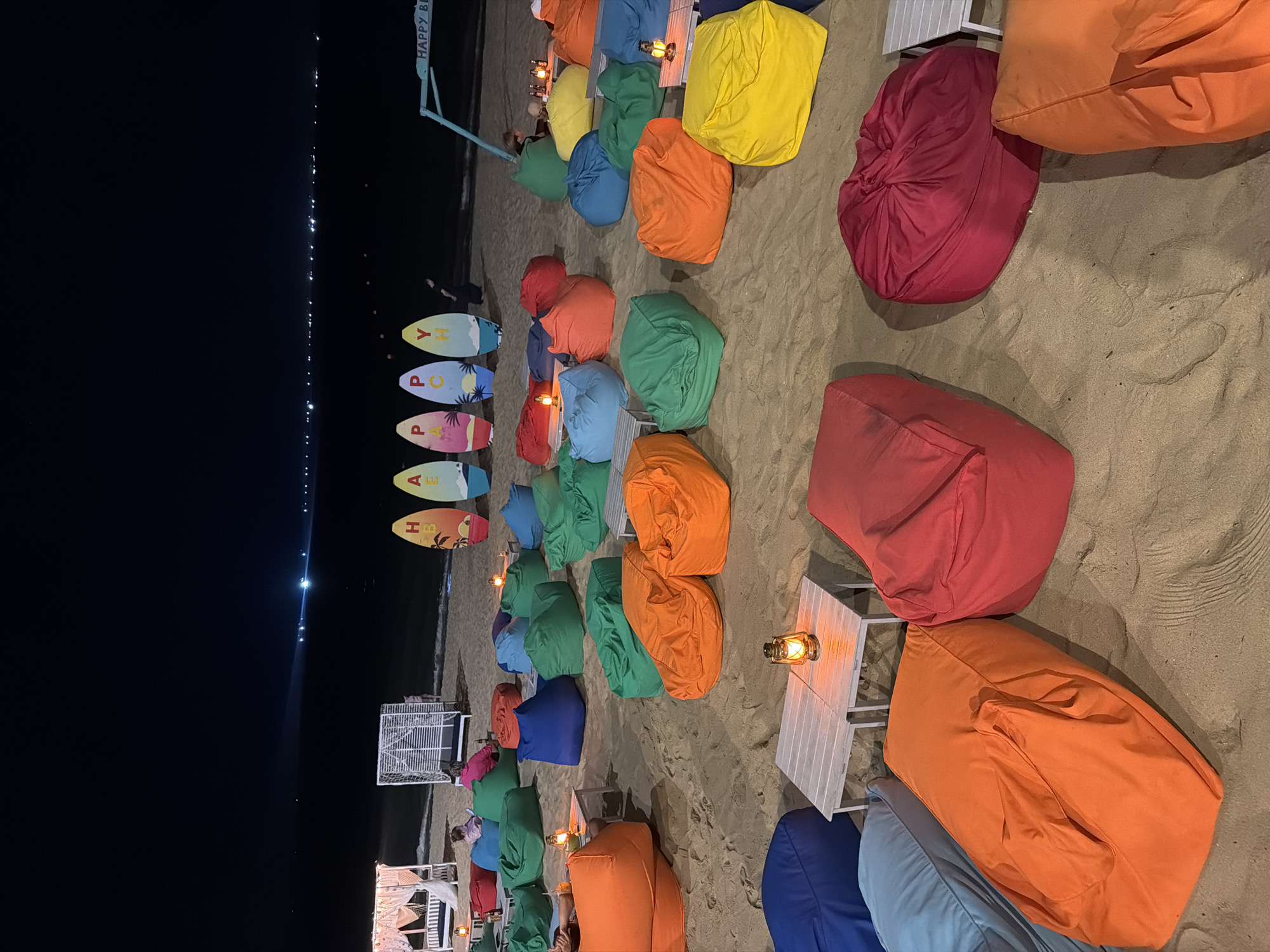 The image depicts a beach scene at night, likely in Nha Trang, Vietnam. Several rows of colorful beanbag chairs (red, orange, green, blue, yellow) are arranged on the sand, each set with a small white table topped with a lit kerosene lamp. In the background, the dark sea is visible, with a string of lights suggesting a distant cityscape along the coast. Five surfboards are standing, with letters that spell "HAPPY". People are barely visible near some lounge setups. The lighting suggests it is evening, with a warm, ambient atmosphere. The surface texture indicates the scene is on a sandy beach.
