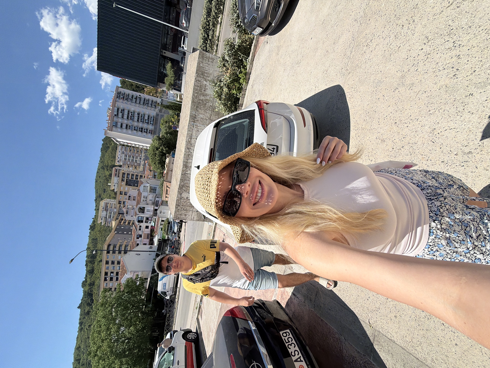 A daytime selfie captures a blonde woman wearing a straw hat and sunglasses, standing on a concrete surface. Behind her and to the side is a male, also wearing sunglasses and a cap, with a yellow and white Puma T-shirt. A black car occupies the bottom right corner with a license plate that is partially visible reading "AS 359." In the background, there are cars parked along the street, with buildings and a green hillside rising in the distance. The sky is a clear blue with some white clouds. The location appears to be in a section of Budva, Montenegro, due to the architectural style of the buildings and the landscape.