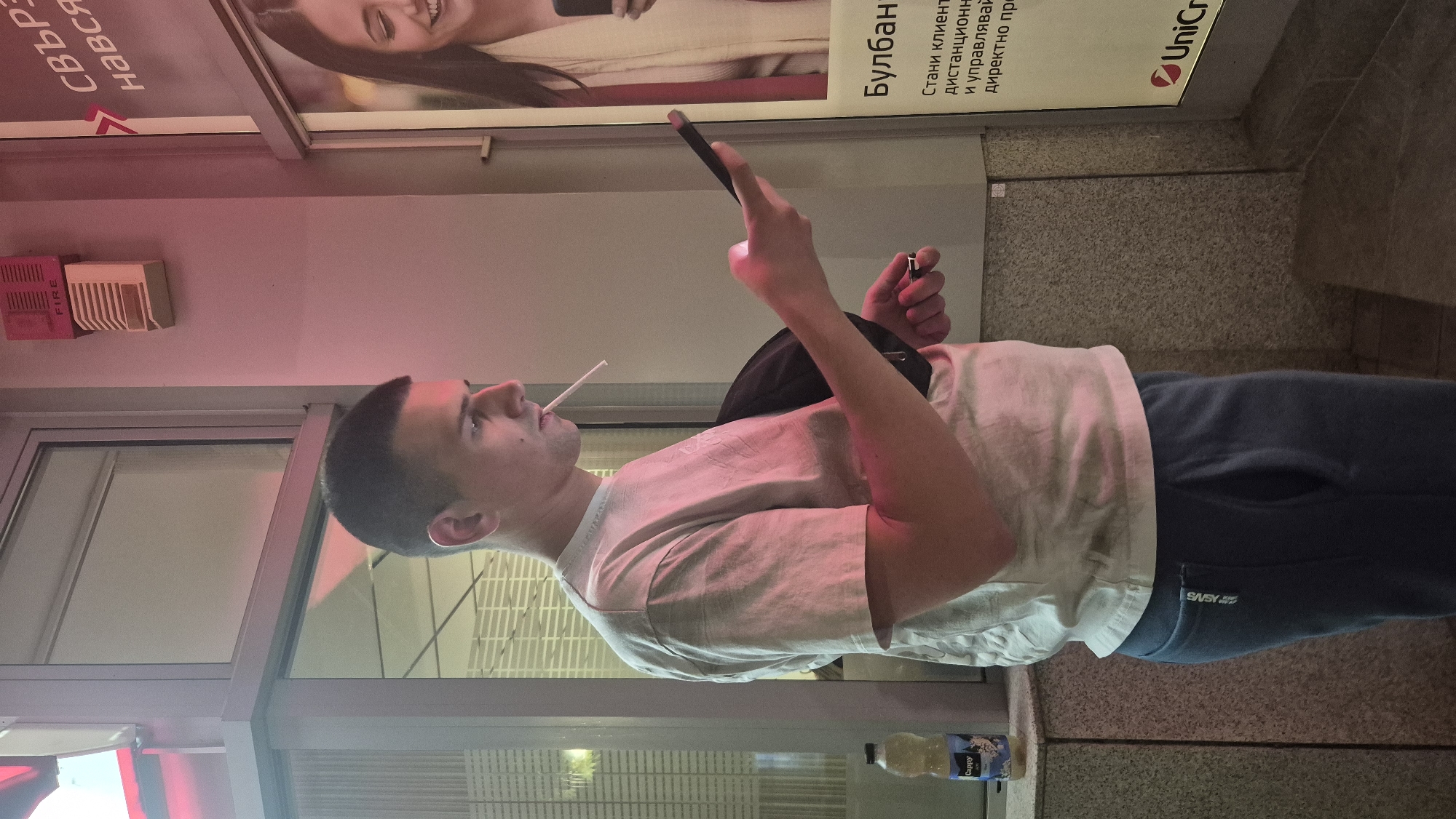 A young man with short dark hair is standing outdoors at dusk, looking at his smartphone. He is wearing a light-colored t-shirt and dark blue sweatpants. A cigarette is held in his mouth, unlit. In his right hand, he is holding the smartphone, and in his left hand, he is holding a small metallic object, possibly a lighter. Behind him, to the left, is a store window with interior lighting. To the right, a billboard displays an advertisement for UniCredit bank. The overall scene appears to be in an urban environment, possibly near a bank branch in Lovech, Bulgaria. The lighting is dim with a pinkish hue, suggesting artificial light sources and the approach of night.