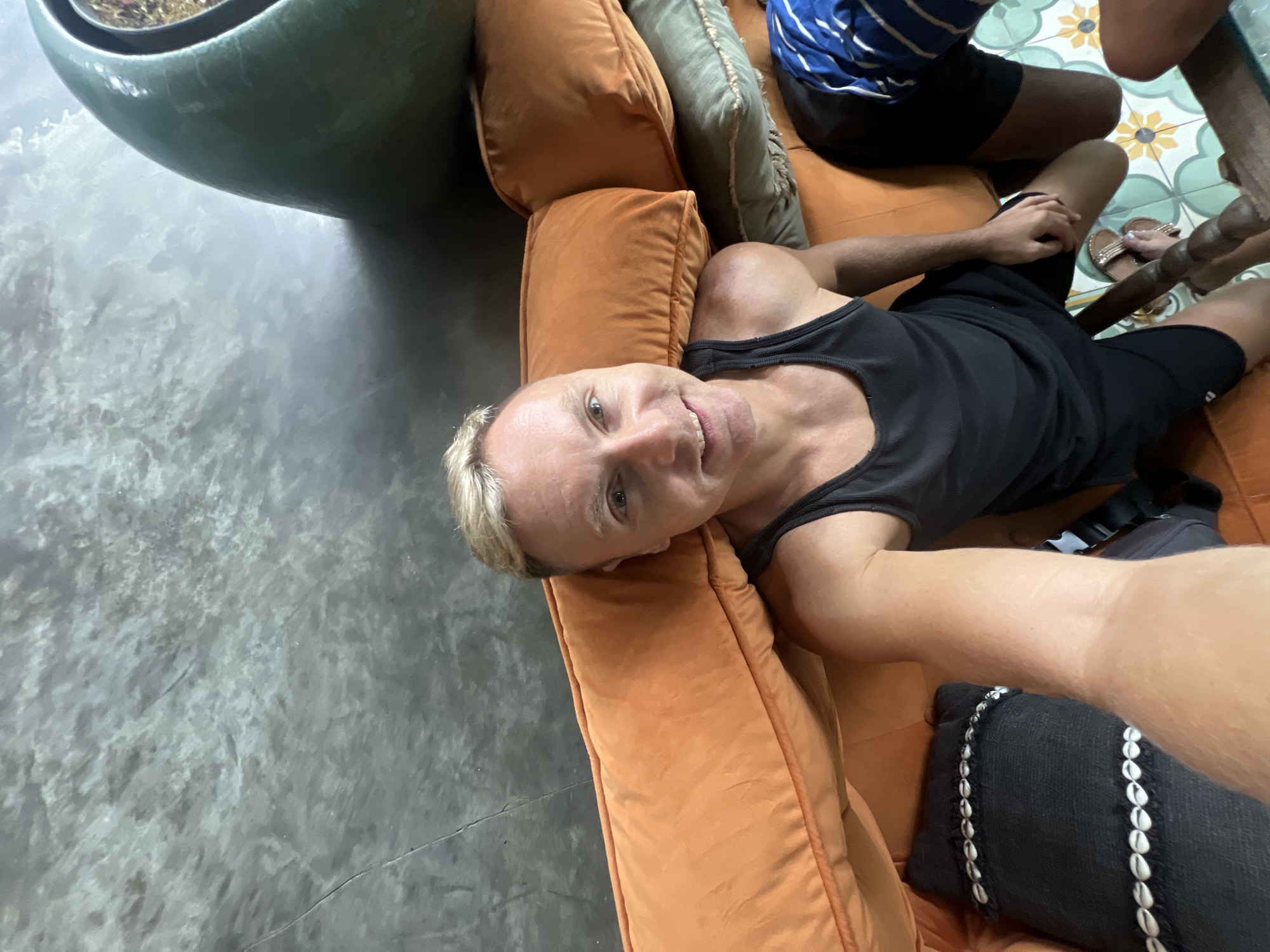 A man with short blonde hair is taking a selfie while sitting on an orange couch. He is wearing a black tank top and black shorts. The couch has several orange and green pillows. To the right of the couch, a large green decorative object is visible. The background is a grey, textured wall. The floor is tiled with a floral pattern. Another person, wearing a blue and white striped shirt and black shorts, is partially visible on the right side of the image. The man's feet are visible, wearing sandals. The scene appears to be indoors, possibly in a living room or lounge area, in Abang, Indonesia.