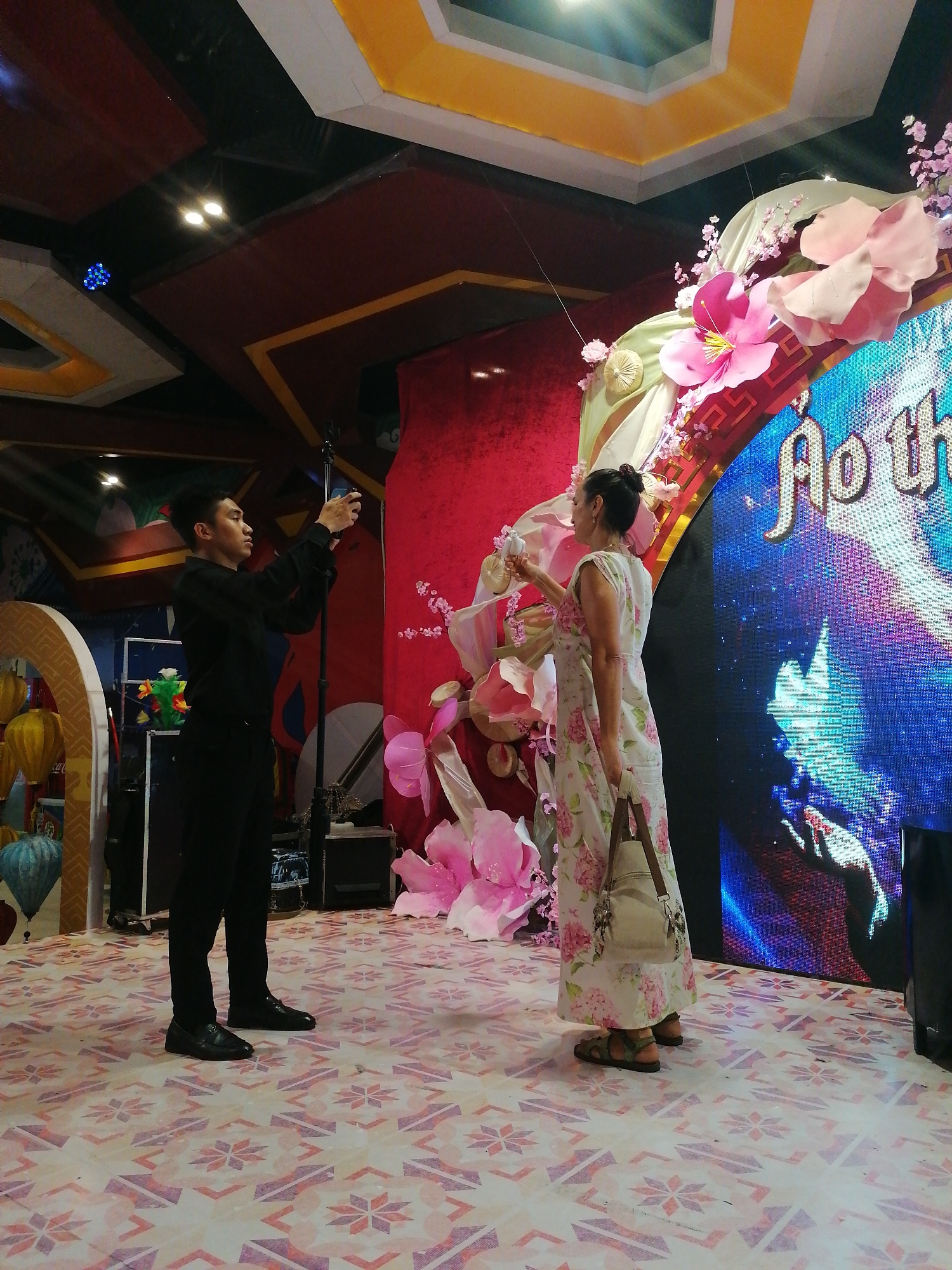 The photograph shows two people on a stage with a patterned floor. A man in a black shirt and pants stands on the left, holding a smartphone in front of him, seemingly taking a picture. To his right, a woman in a light-colored floral dress stands partially facing him with a white object in her raised right hand and a light-colored handbag hanging from her left shoulder. Behind the woman is a decorated archway with pink flowers and a video screen displaying the words "Ao th" in a decorative font. The background includes red draping and additional decorations. The scene appears to be an event or performance setting.