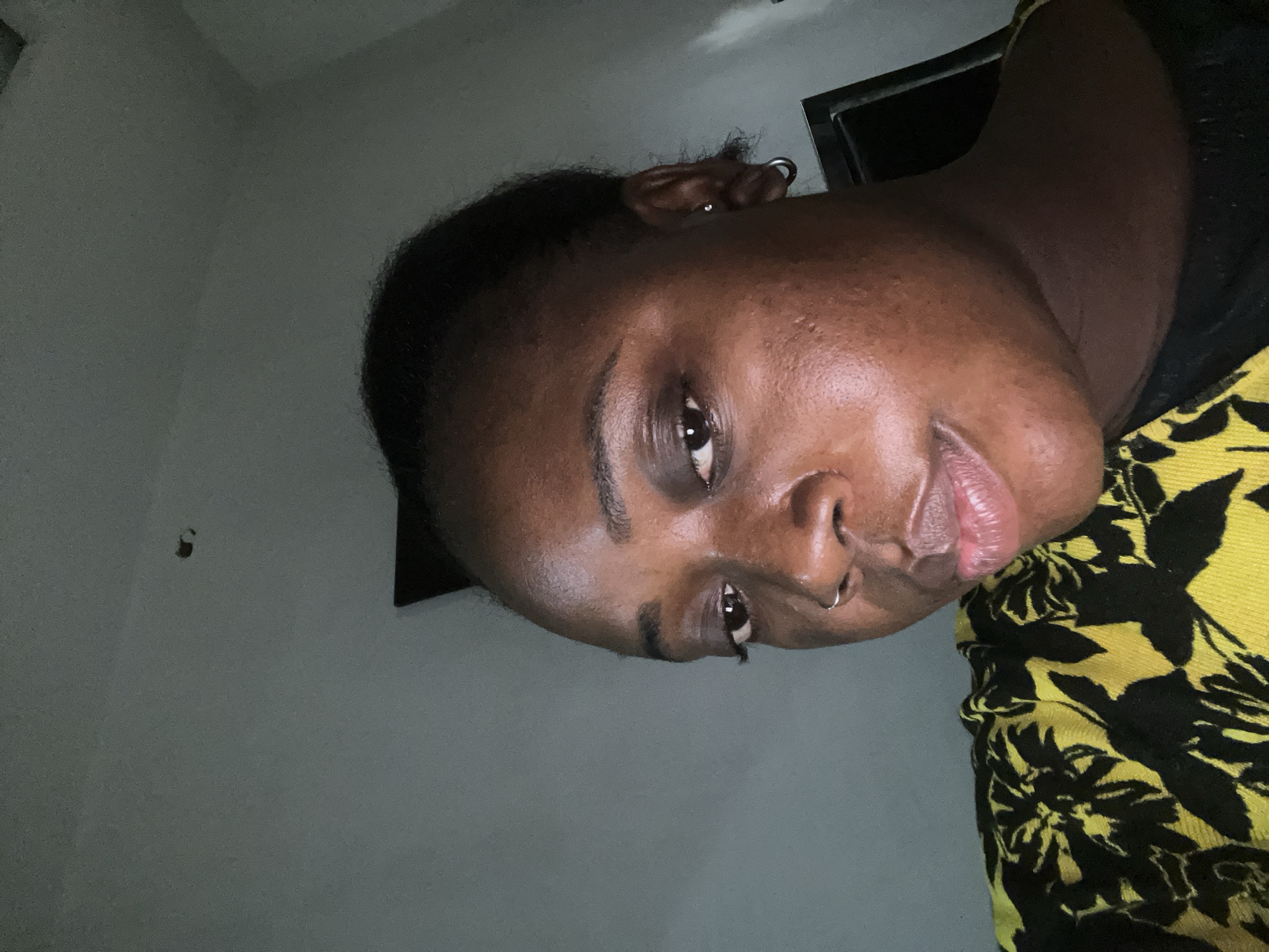The image is a close-up, low-angle selfie of a young Black woman indoors, likely in a residential setting in Jos, Nigeria. She is looking slightly off-camera with a neutral expression. Her skin appears smooth with some natural texture. She has dark eyebrows, dark eyes, and full lips with a subtle sheen. She is wearing a silver hoop earring in her left ear and a small hoop nose ring. Her hair is pulled back neatly. She is wearing a black shirt with a vibrant yellow and black floral pattern on the shoulder and upper chest area. The background is a plain, light-colored wall with a visible imperfection, possibly a small hole or discoloration, on the upper right side. To the right of the woman's head, a dark rectangular object, possibly a door or a piece of furniture, is partially visible. The lighting is dim, creating shadows that highlight the contours of her face. The overall environment suggests a casual, indoor setting.