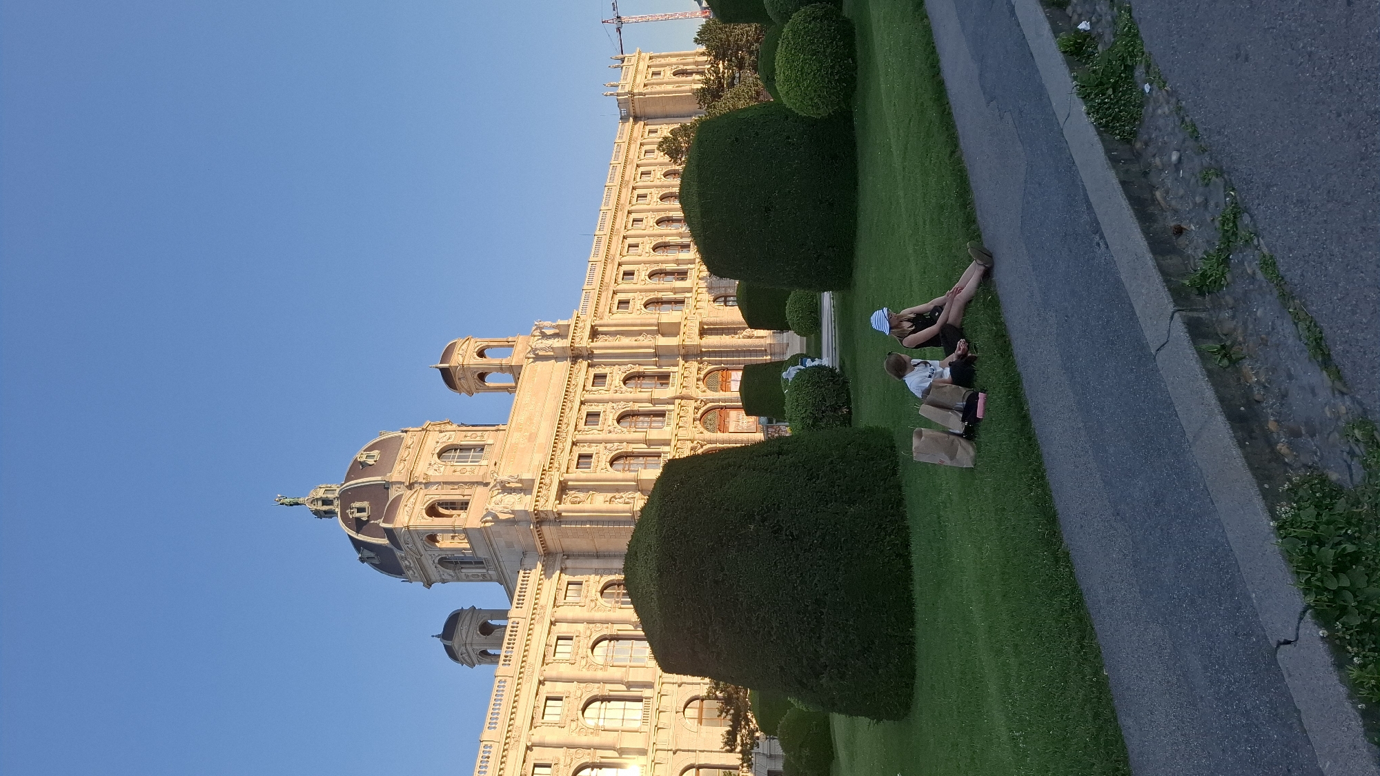 The image shows the Naturhistorisches Museum in Vienna, Austria, bathed in the warm light of what appears to be late afternoon or early evening. Two young individuals are sitting on the lawn in front of the museum, with their legs extended towards a paved path. The person on the left is wearing a white t-shirt and dark shorts, while the person on the right is wearing a dark dress and a blue and white striped hat. They are positioned next to several brown paper bags, suggesting they may have been shopping or picnicking. The museum itself is a grand, ornate building with a large dome and intricate architectural details, set against a clear blue sky. The foreground is dominated by a neatly manicured lawn with several large, cone-shaped bushes and smaller, rounded hedges. The overall scene conveys a sense of peaceful relaxation in an urban, historical setting.