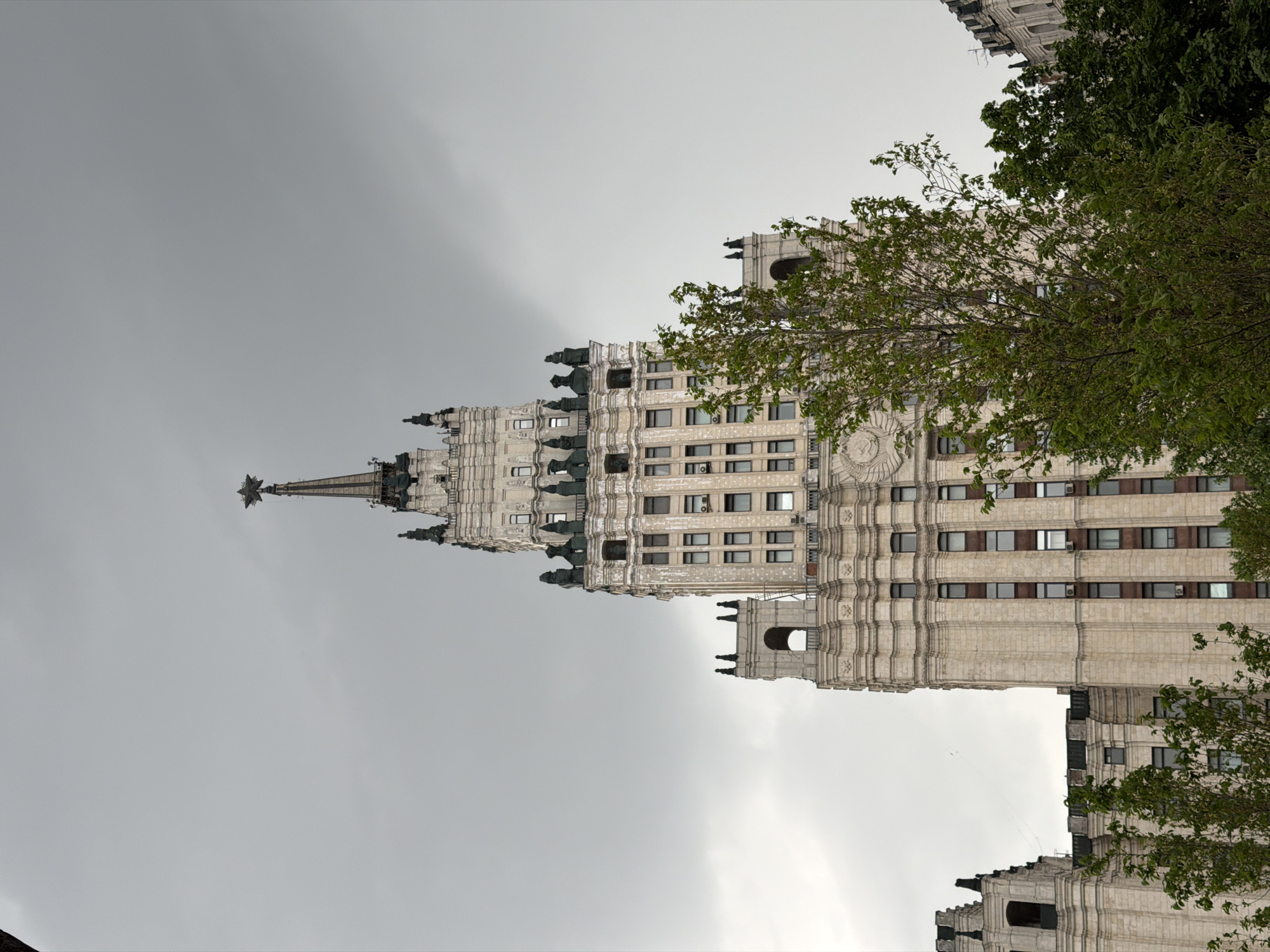 The image shows a building in Moscow, Russia known as one of the "Seven Sisters." This is a group of skyscrapers constructed in the Stalinist style. The building is captured from a low angle, emphasizing its height and intricate architectural details. The facade is predominantly beige or light grey, with symmetrical rows of windows and ornate decorations. The upper part of the building tapers into a spire, topped with a star. The sky above is overcast, creating a muted, grey background. Trees with green leaves are visible in the foreground.