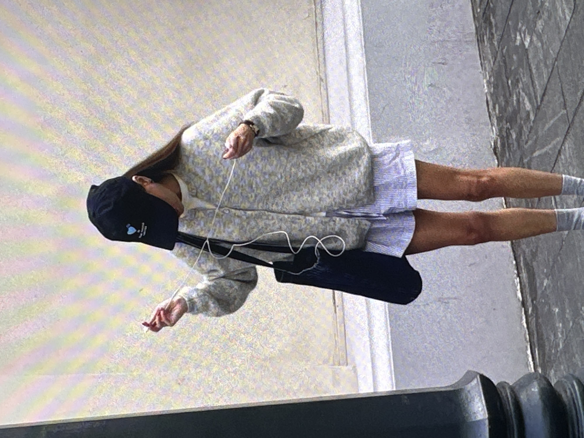 A person is walking on a sidewalk in Yerevan, Armenia, near a building with a concrete façade and a gray brick walkway. The person, likely female, wears a black baseball cap, a light-colored, patterned sweater over a striped short dress, and gray socks. They are carrying a dark-colored shoulder bag. The person is holding white earbuds and appears to be listening to music. A gray cast iron bollard is visible in the lower portion of the photo.