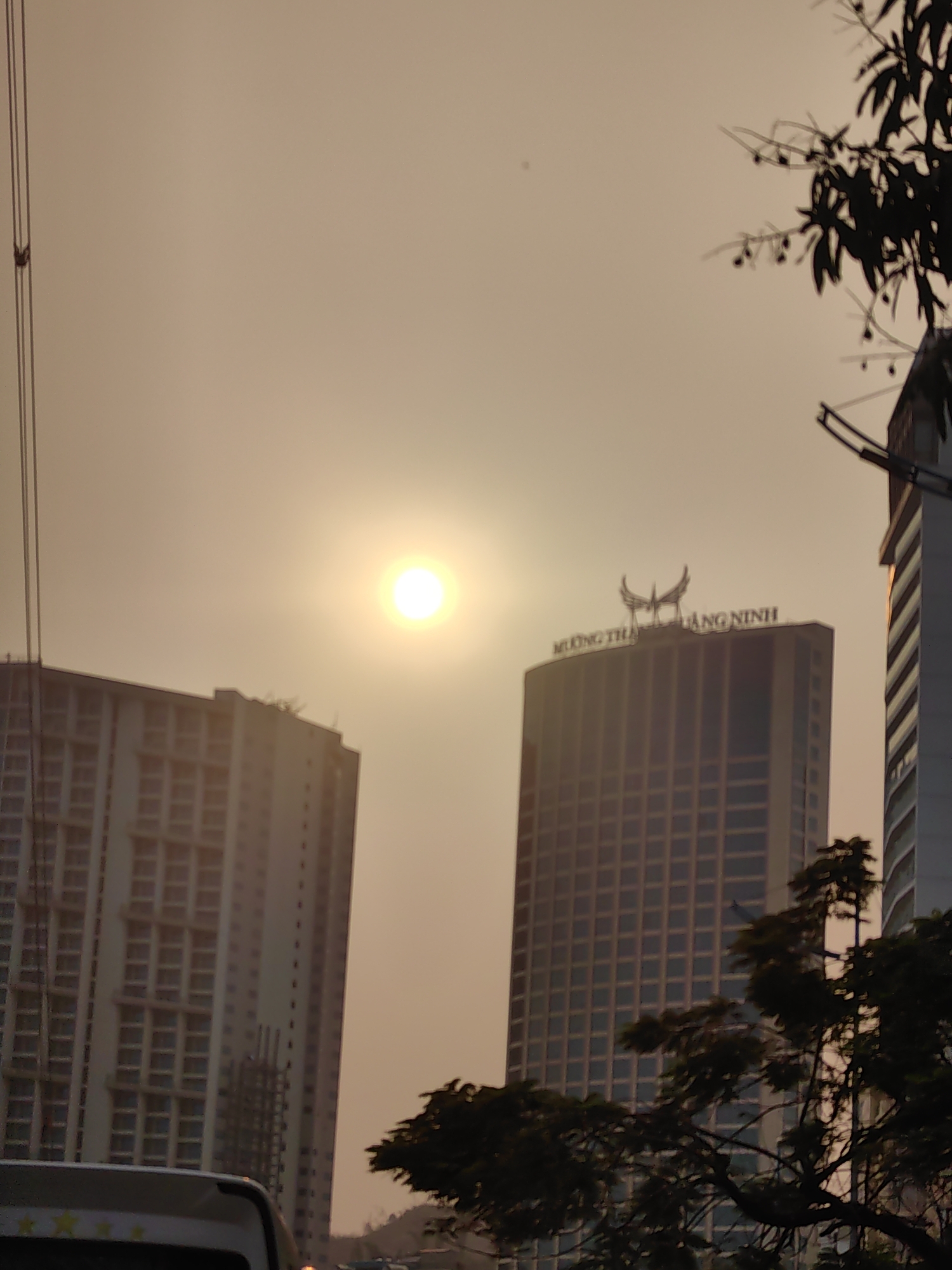 The image depicts a cityscape at sunset in Bai Chay Ward, Vietnam. Two tall buildings dominate the view: one on the left with a simple rectangular structure and multiple windows, and the other on the right with a distinctive ornamental structure on top reading "MUONG THANH QUANG NINH". The sun is positioned between the buildings, casting a bright glow. Trees are silhouetted at the bottom of the frame, and a vehicle is partially visible in the lower left corner. Power lines are seen in the upper left of the shot, and another building is on the far right with what appears to be a communications tower. The sky is a muted orange color, indicating a late afternoon or early evening setting.