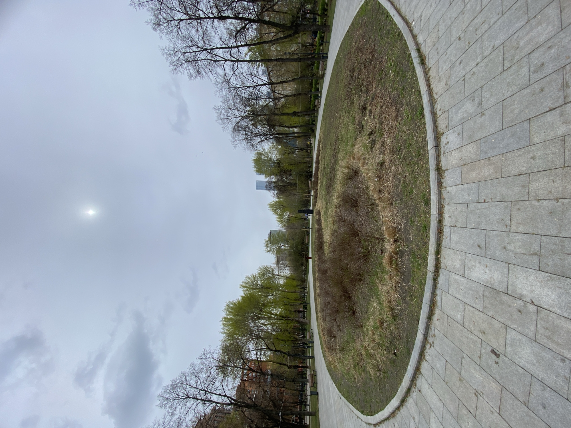 This landscape photograph presents a serene urban park scene under an overcast sky. A wide, curving pathway made of grey rectangular pavers originates in the bottom right foreground and arcs gracefully towards the upper left, delineating a large, somewhat oval-shaped central ground area. This central section features a mix of dry, brownish grass and emerging green patches, along with sparse, dark, shrub-like vegetation.
Numerous trees line both sides of the park. Those closer to the viewer, particularly on the left side of the frame, are mostly bare-branched, while trees further in the background display vibrant new green leaves, indicating an early spring transition. In the distance, beyond the treeline, several tall, modern city buildings are visible, confirming the park's urban setting. A few small, indistinct human figures can be seen walking along the pathway further away.
The sky is uniformly covered with grey clouds, with the sun appearing as a bright, hazy glow in the upper left quadrant, trying to penetrate the cloud cover. The time of day is daytime, but the overcast conditions create soft, diffuse lighting. The blend of bare trees and budding foliage, along with patchy grass, strongly suggests the season is early spring. The scene evokes a quiet, possibly cool atmosphere in a public park, consistent with the described location of Moscow, Russia. No visible text is present in the image.