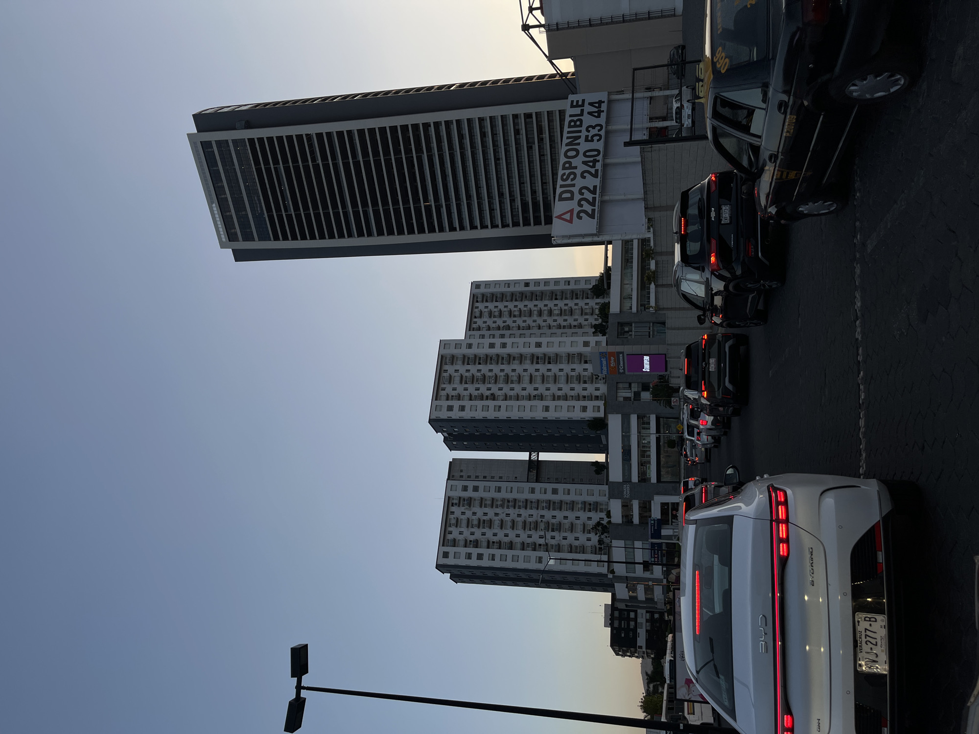 The image displays a busy urban street scene in San Bernardino Tlaxcalancingo, Mexico, observed during what appears to be late afternoon or early evening under a clear sky. The setting is characterized by several prominent buildings and a line of vehicles moving along the street.
In the background, a tall, modern skyscraper with numerous horizontal window panels or louvers stands prominently. A large billboard attached to its side reads "▲ DISPONIBLE 222 240 53 44", indicating property availability and a contact number. Below and around this building, several other multi-story structures, possibly residential or mixed-use, are visible, featuring numerous windows and balconies.
The foreground and midground are dominated by traffic. The rear of a white BYD electric sedan, with its distinctive full-width LED taillights and "BYD" and "BYDKING" branding, is clearly visible. Its license plate, "VERACRUZ YVJ-277-B", identifies its registration. Behind it, a dark Chevrolet SUV and other cars are queuing. Further back in the traffic, a black taxi can be seen, partially displaying "990", "PZ0709", and "ENV-173-C" on its body. A street light is visible in the lower left of the frame.
Various commercial signs are scattered throughout the urban landscape, including "Walmart", "Cinemex", "SONATA TOWERS", "STARTUPERO", "Pickup", "art", and "IN". The scene captures the typical atmosphere of a metropolitan area with ongoing vehicular activity and diverse architecture.
