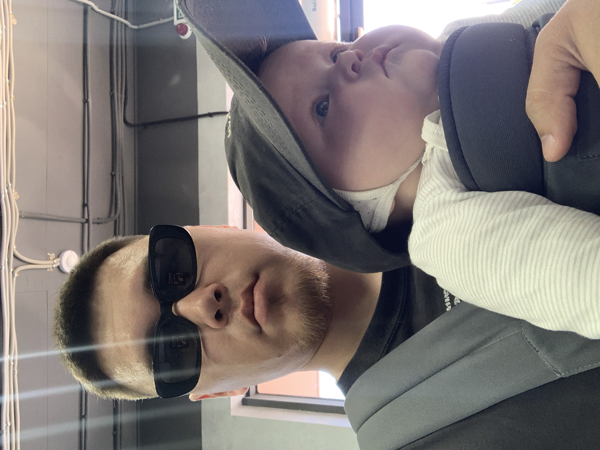 A man with short brown hair and a light beard, wearing black sunglasses and a black t-shirt, is pictured from the chest up, holding a baby in a carrier. The baby, wearing a white and grey striped onesie and a dark baseball cap that shades its face, is positioned in front of the man. The man's left arm is visible, supporting the carrier. The background shows an indoor environment with ceiling lights and electrical conduits. The specific location in Stavropol, Russia, is not identifiable from the image.