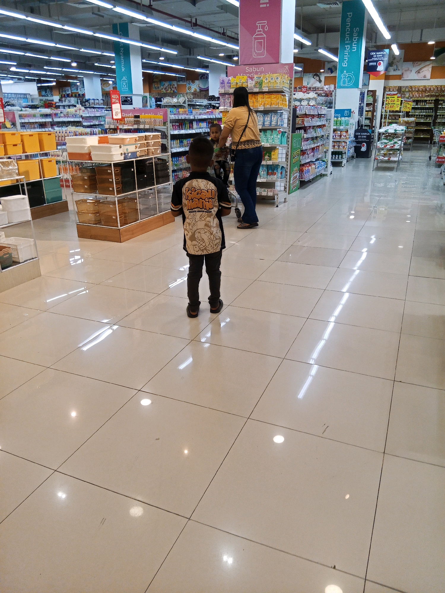 The image depicts an interior view of a well-lit supermarket in Lubuklinggau, Indonesia. In the foreground, a young boy wearing a dark shirt with a graphic print and dark pants stands with his back to the camera, facing further into the store. To his left and slightly behind him, another child is partially visible. A woman in a yellow shirt and dark pants is pushing a shopping cart, her back also turned towards the camera, positioned to the right of the boy. The supermarket aisle is lined with shelves stocked with various products, including cleaning supplies and household goods. The floor is tiled in a light beige color, reflecting the overhead fluorescent lighting. Signs with Indonesian text are visible on the shelves and hanging from the ceiling, indicating product categories and prices. The overall environment suggests a typical shopping experience in a modern retail space.