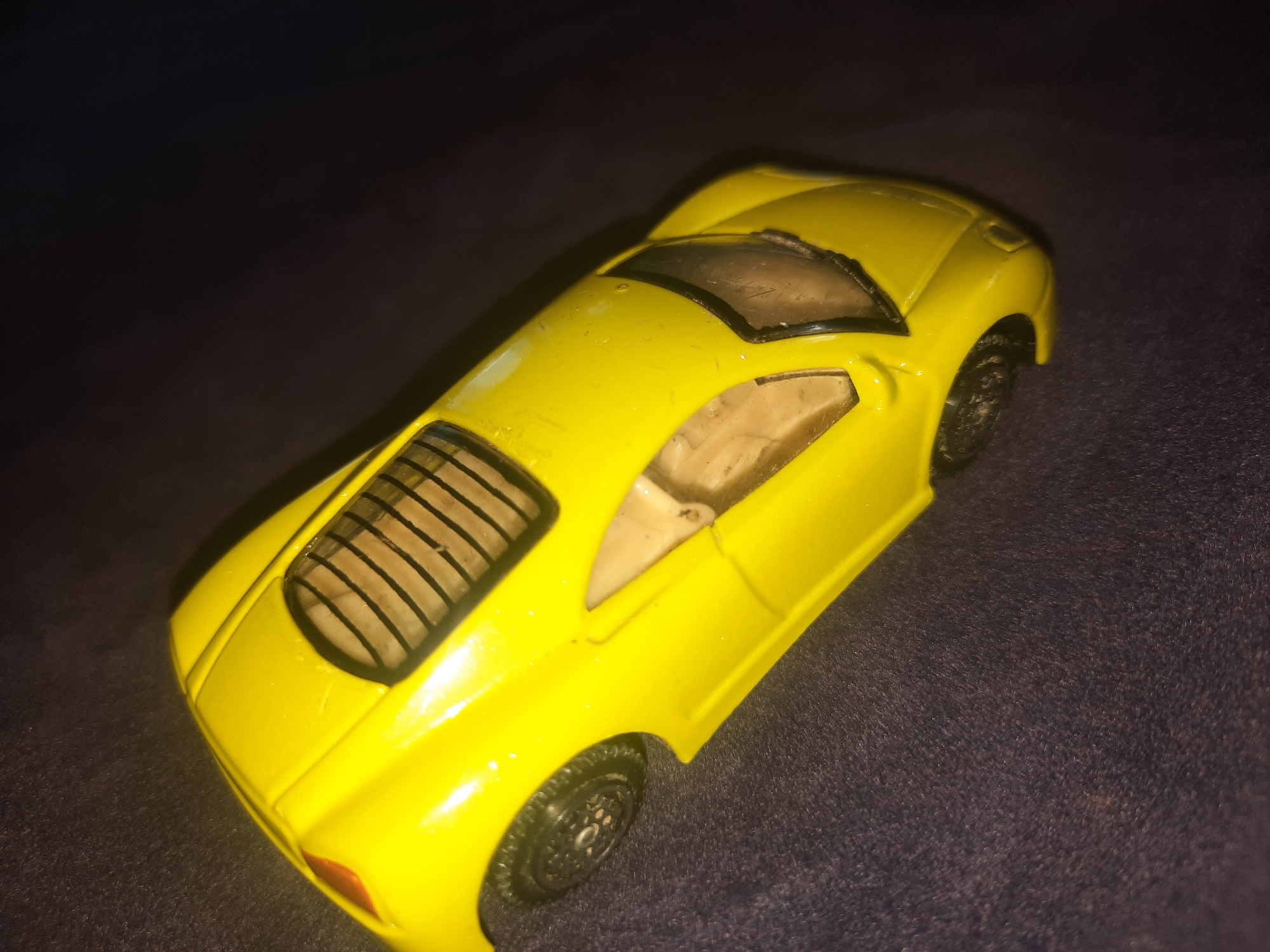 A bright yellow toy sports car is photographed at an angle against a dark, textured surface. The car has a tinted windshield and windows, and a rear engine cover with horizontal slats. The background is dark and out of focus, suggesting dim lighting conditions. There are no people or any identifiable landmarks of the city of Шклоў, Belarus visible in the image.