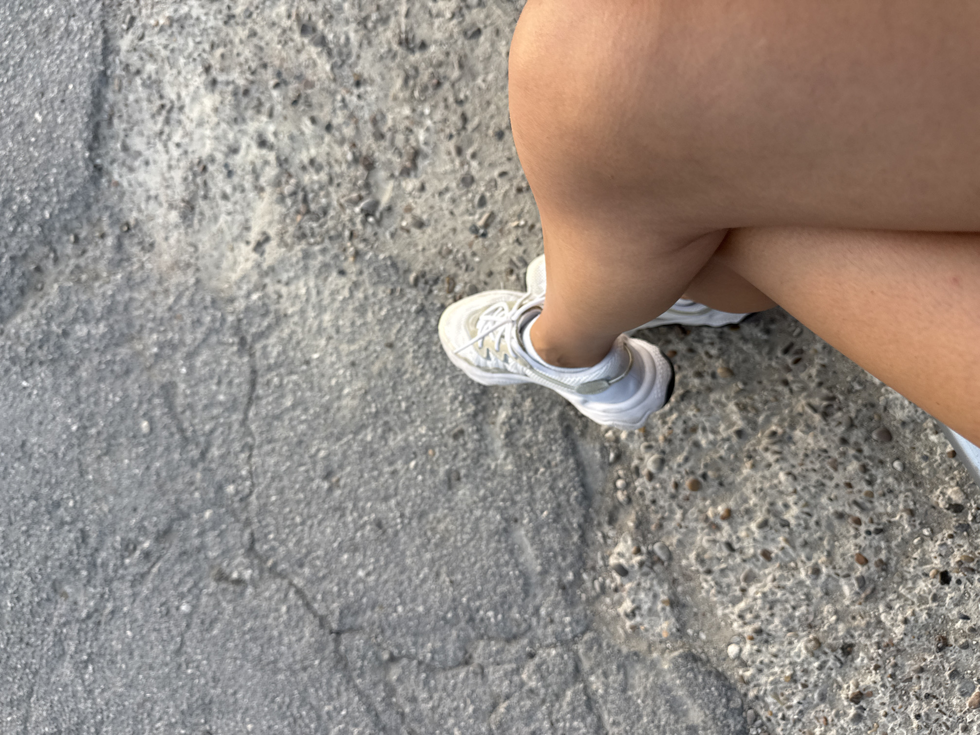 The image shows a close-up, high-angle view of a person's legs and feet, wearing white athletic shoes, crossed at the knees. The ground beneath is a cracked asphalt surface with visible pebbles and debris. The lighting suggests it is daytime, with shadows indicating the sun's position. The person's skin tone is tan. The environment appears to be an outdoor urban setting, possibly a sidewalk or road, in Cimișlia, Moldova.