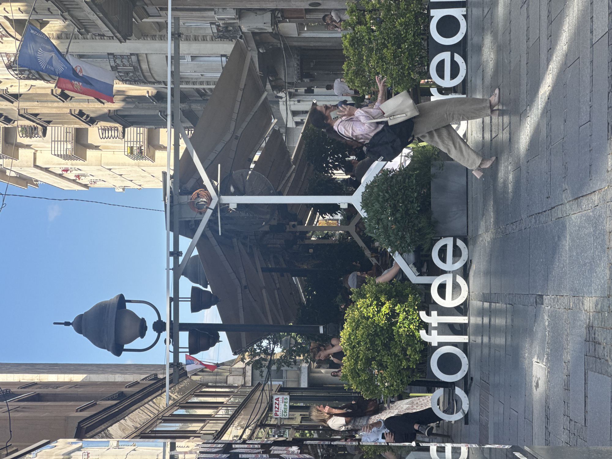 This image depicts a street scene in Belgrade, Serbia, likely in a pedestrian zone or a popular cafe area. The foreground features large, white, three-dimensional letters spelling out "coffee," with another partial word starting with "rea" visible to the right. The letters are placed on the cobblestone pavement.
In the midground, a woman in light-colored pants and a pinkish top is walking towards the right side of the frame, carrying a black backpack and a light-colored tote bag. She appears to be speaking on her phone. Behind her, there are several people seated at tables outside a cafe, surrounded by lush green bushes. The cafe has a retractable awning providing shade.
The background reveals the architecture of Belgrade, with multi-story buildings featuring balconies and windows. Flags, including the Serbian flag (red, blue, and white tricolor) and another flag with a blue and white design, are visible hanging from one of the buildings. A vintage-style street lamp with a dark-colored shade is also prominently featured on the left side of the image. The sky is clear and blue, indicating daytime. A sign for "Pizza Crepes" is visible on a building to the left. The overall atmosphere suggests a vibrant, urban setting with outdoor dining and pedestrian activity.