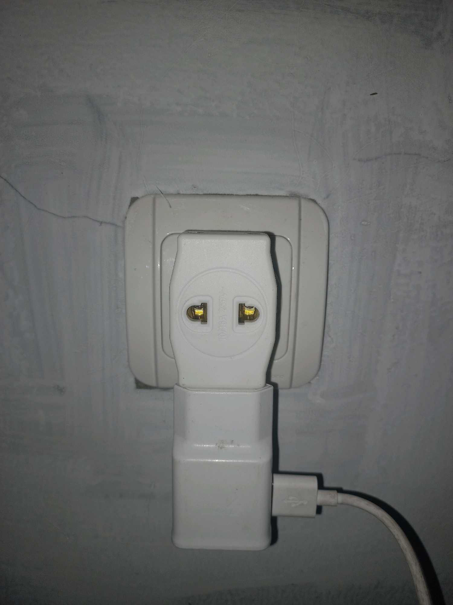 The image shows a close-up of a white electrical outlet adapter plugged into a wall socket. A white USB charger is also plugged into the adapter, with a white USB cable extending to the right. The wall appears to be concrete or stucco, with some visible cracks and a textured surface. The lighting suggests an indoor environment, possibly a domestic setting. The location is identified as Gambiranom, Indonesia.
