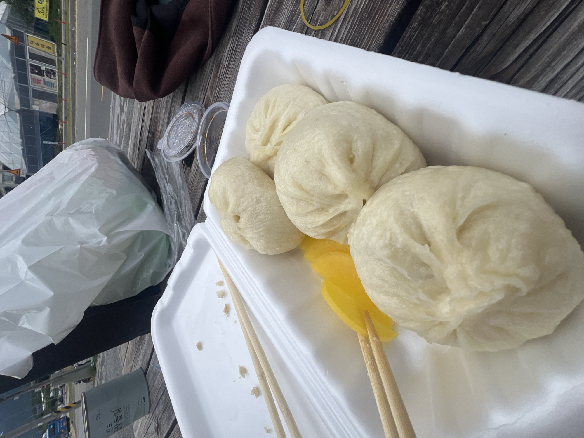 At a street-side picnic table in Dongmyeong-myeon, South Korea, a white styrofoam container holds a meal of steamed buns and yellow pickled radish. Two pairs of wooden chopsticks rest on the container, one pair piercing the pickled radish. Nearby, a white plastic bag, a brown jacket, and two small plastic dipping sauce containers are also visible. In the background, a road runs past several commercial buildings with signage in Korean.