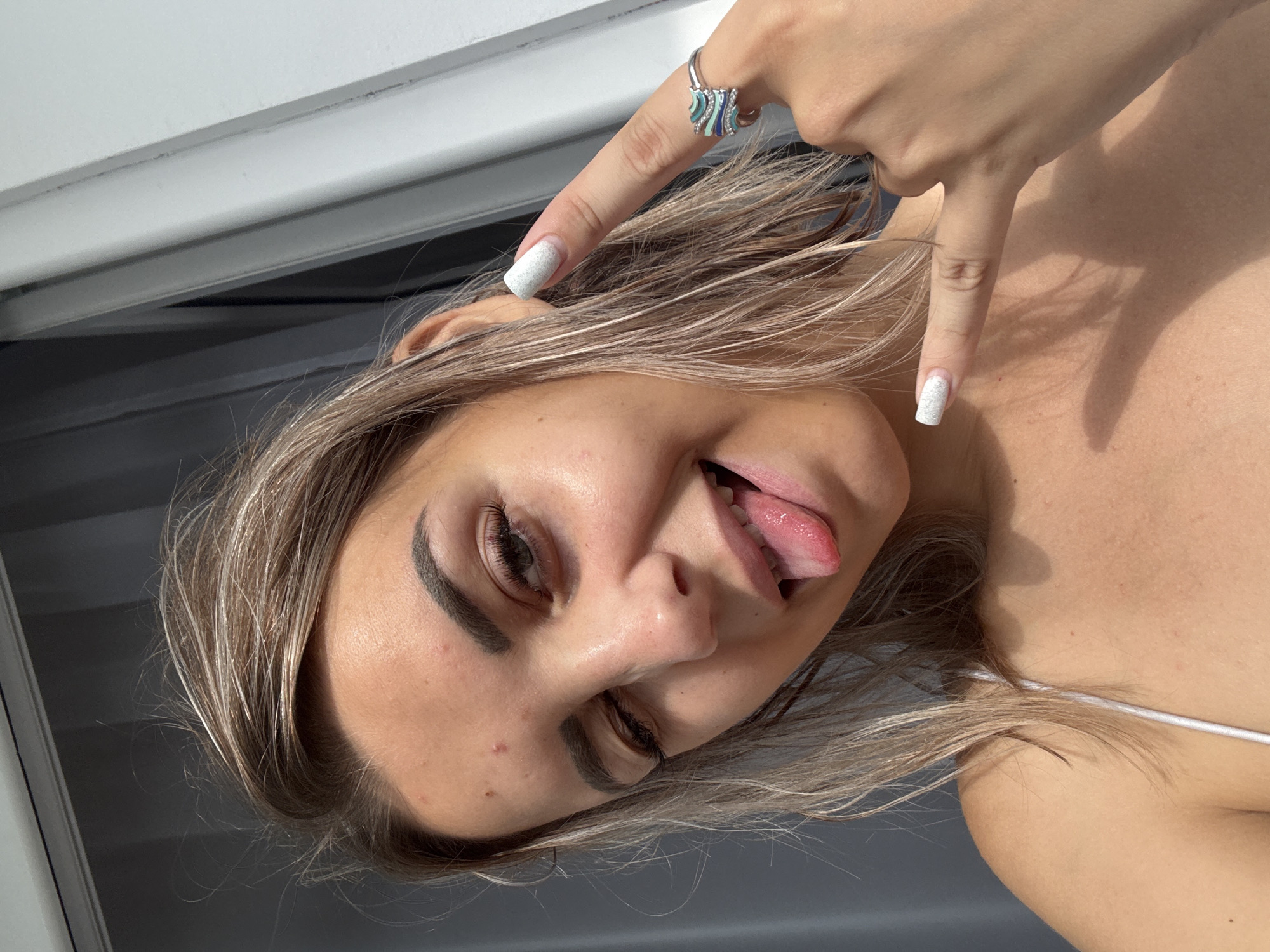 In this close-up, medium shot, a young woman with blonde hair is pictured outdoors. She is looking towards the camera with her right eye closed in a wink. Her tongue is sticking out to the left, and she is wearing a white top. On her right hand, she is making a rock-and-roll gesture with her index and pinky fingers extended. She is also wearing a ring on her right ring finger. The background suggests an urban environment with a blurry grey curtain on the left and a white vertical structure on the right. There are no identifiable landmarks to indicate the specific location in Kholmsk, Russia.