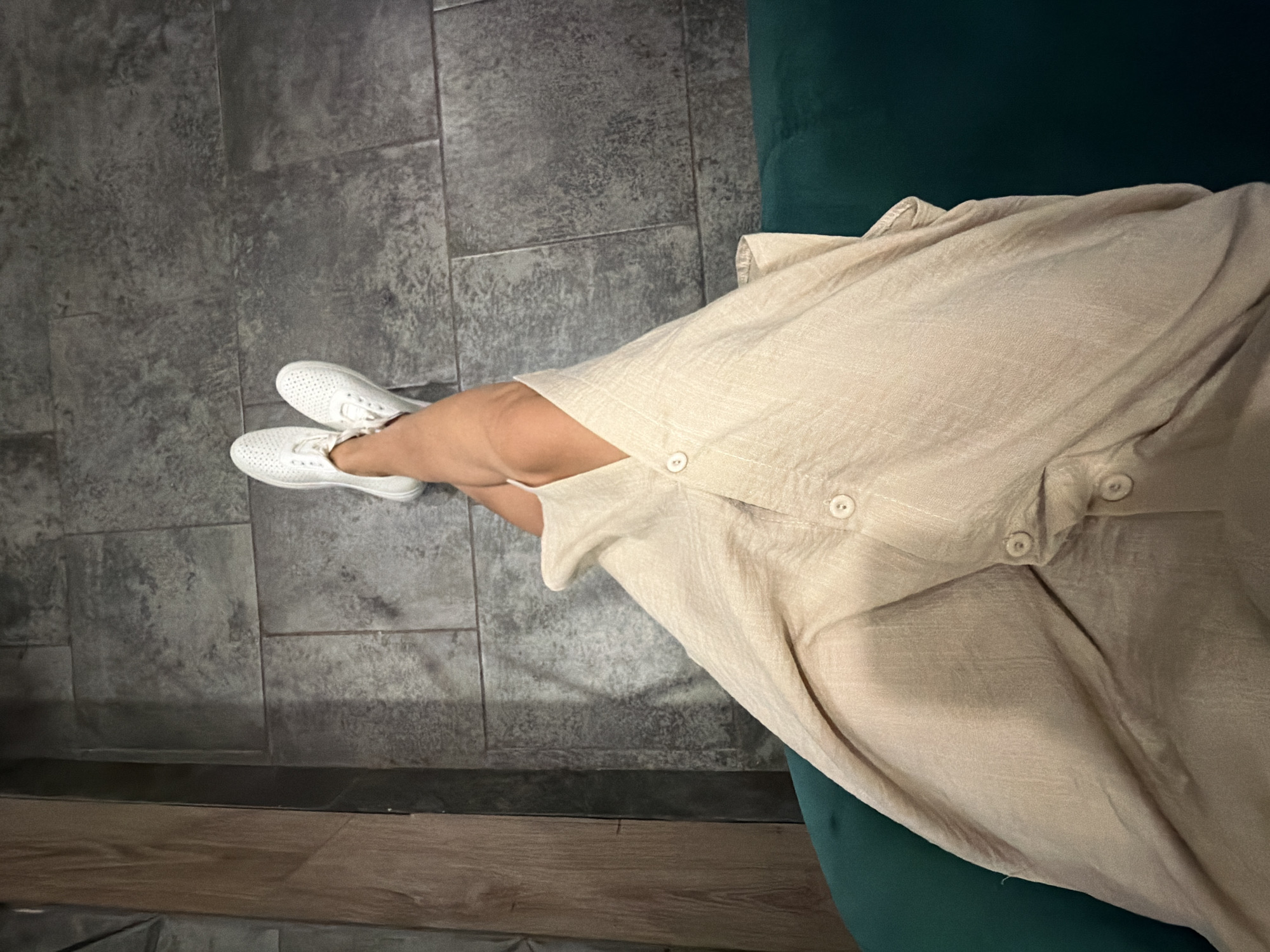 A person is sitting on a dark green sofa, their legs crossed and resting on a tiled floor. They are wearing a light beige dress with buttons down the front and white sneakers. The floor is made of large, dark gray rectangular tiles. To the left of the floor is a wooden wall. The image is taken from a high angle, looking down at the person's legs and dress. The lighting is dim, casting shadows on the floor. There is no indication of the city of Chaikovsky, Russia in the image.