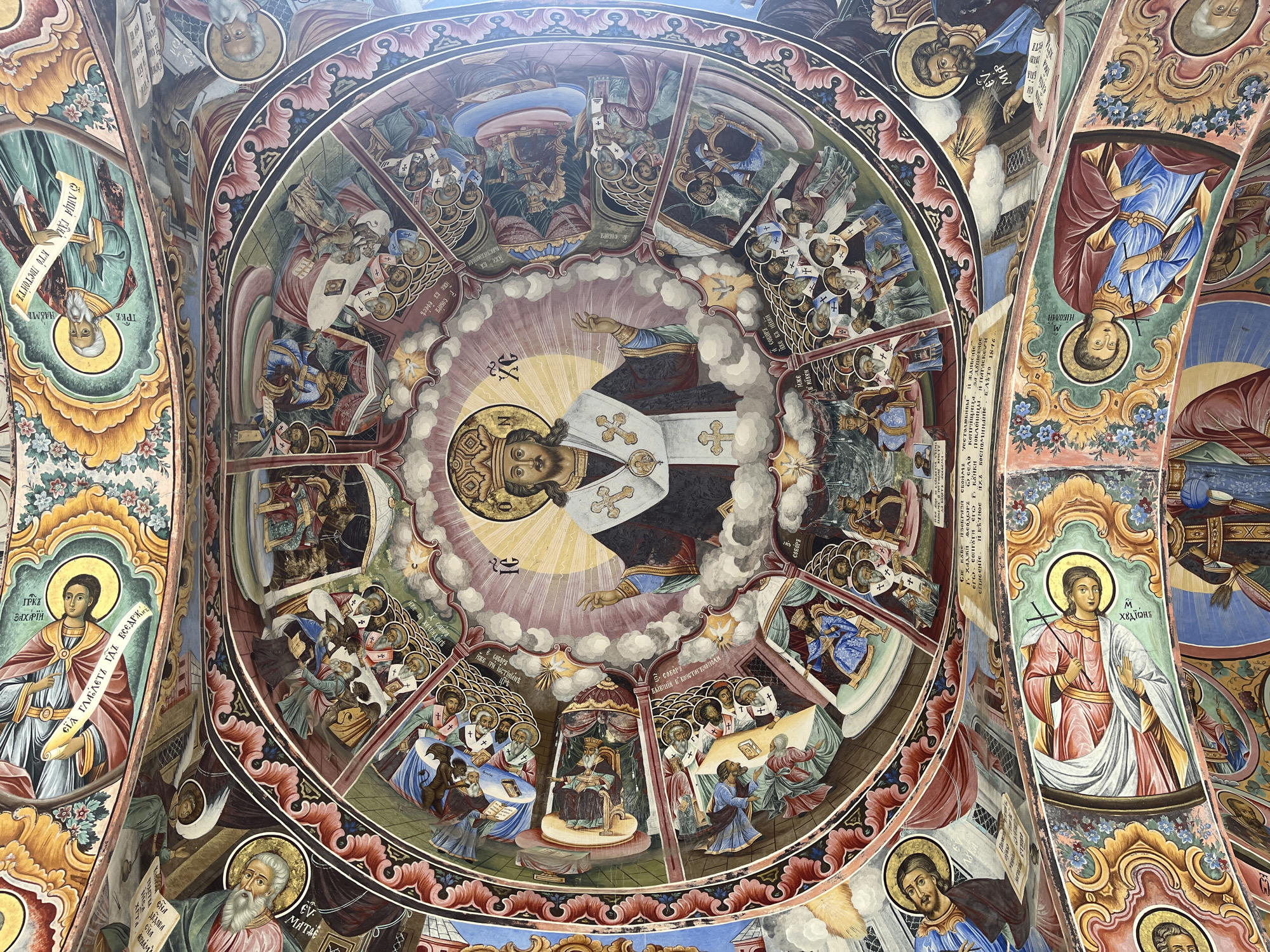 The image shows a ceiling fresco, likely located in the Rila Monastery (Rilski manastir) in Bulgaria. The fresco is a circular composition with a central figure, presumably a religious figure, surrounded by clouds. The figure is dressed in black and white robes. Around the central circle are multiple sections depicting scenes with groups of people, possibly biblical or historical events. Figures have halos above their heads. There are smaller portraits framed by decorative borders. Inscriptions in a non-Latin alphabet are present, including names and possibly dates. The fresco uses a palette of blues, reds, yellows, and browns. The overall style is Byzantine or Eastern Orthodox.