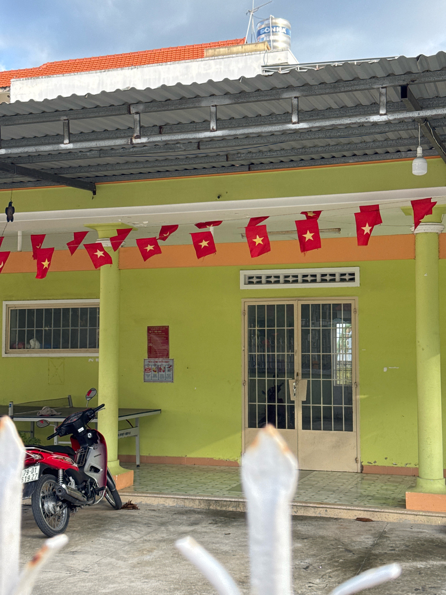 This image captures a storefront or building entrance in Nha Trang, Vietnam, likely a public or commercial establishment given the flags and signage. The building features a bright lime-green facade with an orange trim beneath a corrugated metal awning supported by metal beams. Red and yellow Vietnamese flags are strung across the awning, creating a festive or patriotic display. A set of double doors with a metal grid is centrally located, with a narrow horizontal window above it. To the left of the doors, there is a larger window with a metal grate. A red and silver Honda motorcycle is parked in front of the building. Behind the motorcycle, a ping-pong table is visible. A red sign with Vietnamese text is mounted on the wall to the left of the doors. The ground is paved with patterned tiles, and the sky above is cloudy. The scene suggests a warm climate.