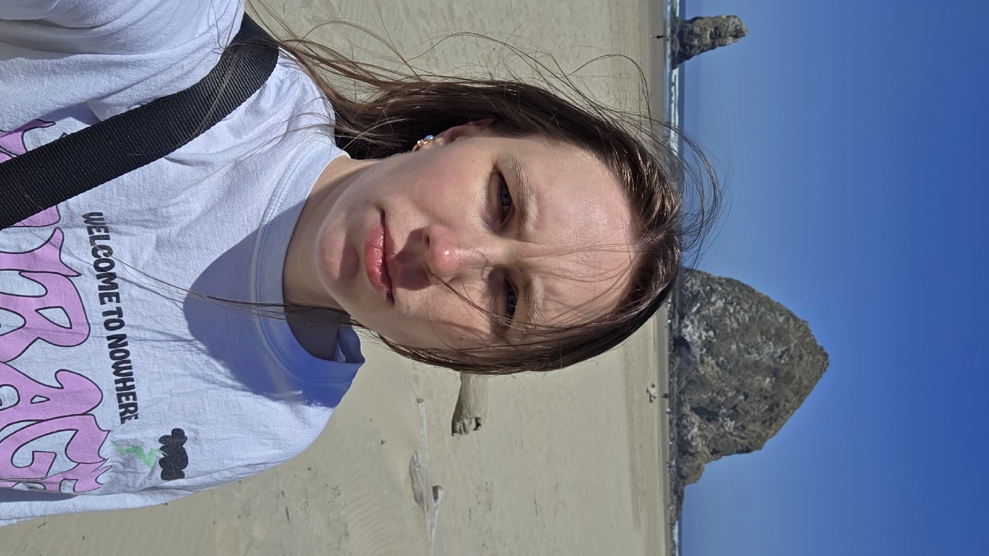 A woman is taking a selfie on a beach in Cannon Beach, Oregon, United States of America. She is wearing a white t-shirt with the text "WELCOME TO NOWHERE" and a graphic of a cloud with lightning. Her hair is blowing in the wind. In the background, the iconic Haystack Rock and a smaller sea stack are visible on the beach. The sky is clear and blue. The sand is light brown, and there are pieces of driftwood scattered on the beach. Several people are visible in the distance on the beach and in the water.