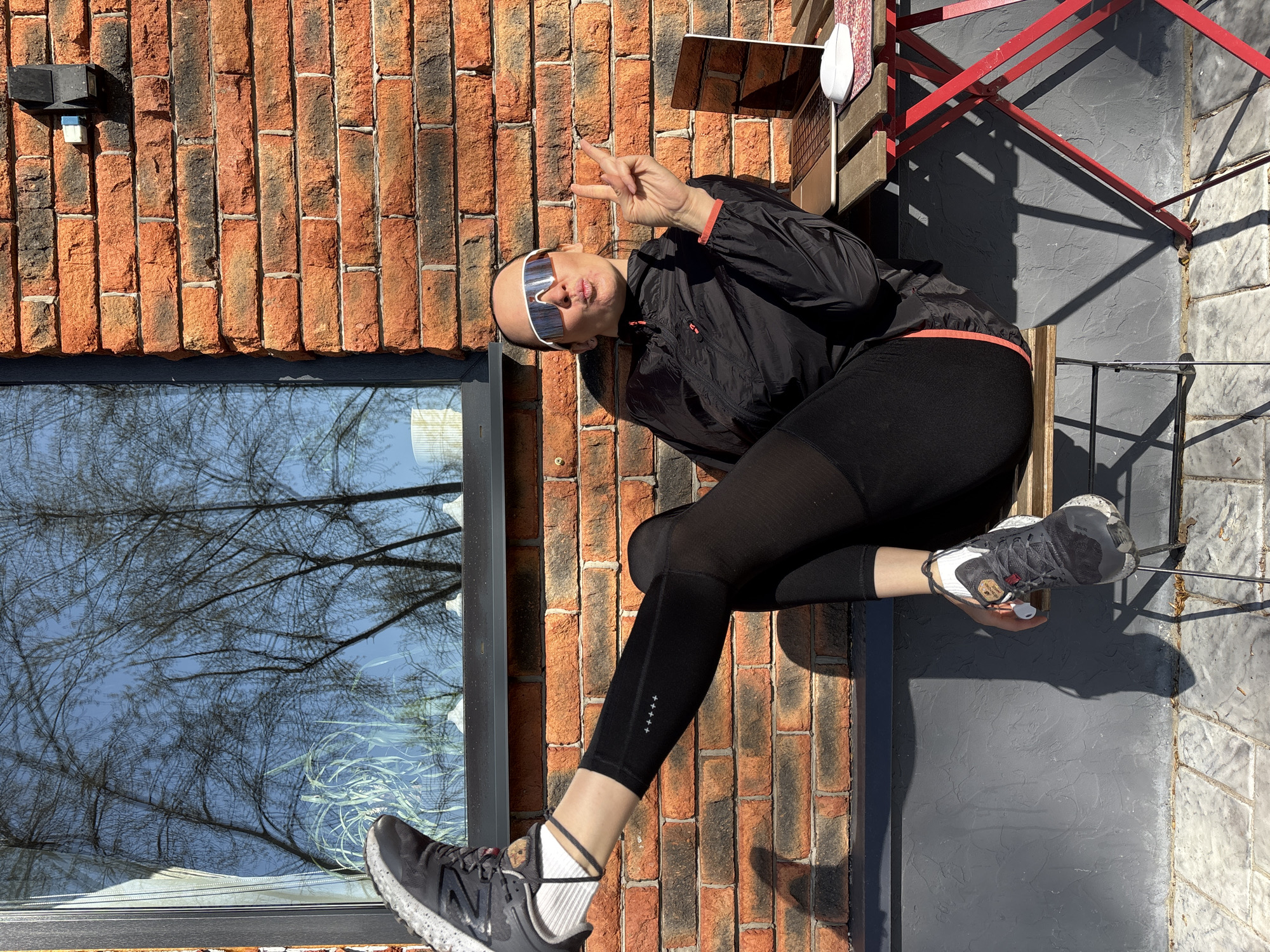 A person wearing sunglasses, a black jacket, and black leggings sits on a red metal chair outside a brick building in Chaikovsky, Russia. The person is giving a peace sign with their right hand. The building has a window reflecting trees and sky. The ground is paved with gray concrete.