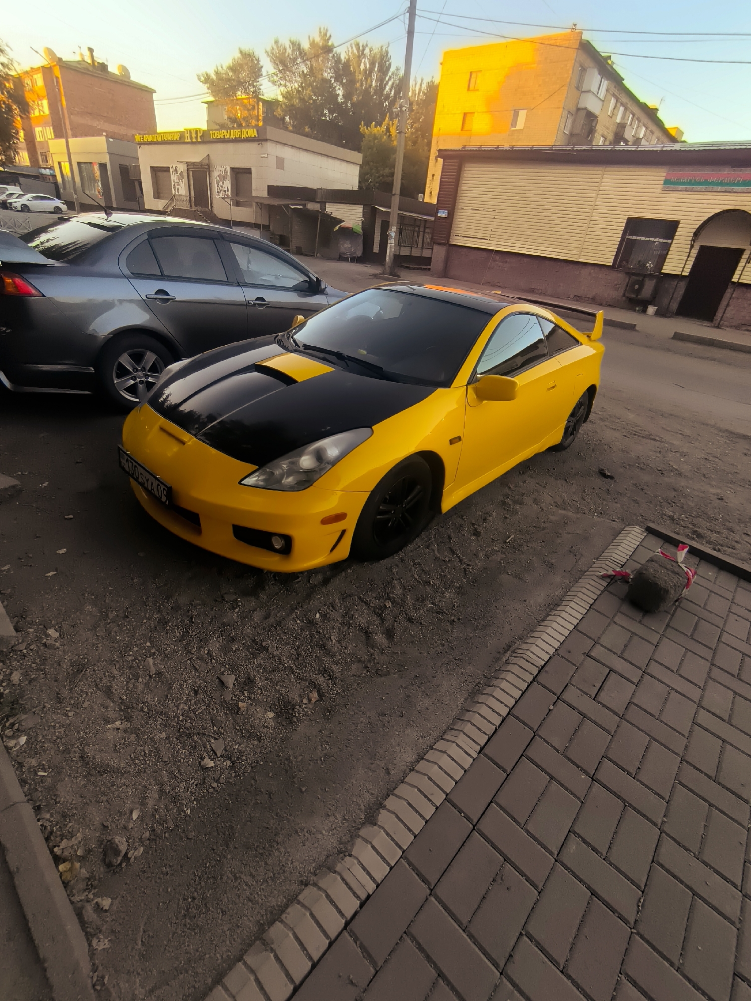 The image shows a yellow Toyota Celica parked on a gravelly surface next to a paved area. In the background, there are buildings, trees, and other cars, suggesting an urban environment. The lighting indicates it is either sunrise or sunset, with warm tones casting a glow on the scene. One of the buildings has signage in Russian that reads "ТОВАРЫ ДЛЯ ДОМА" (Goods for the home). The license plate on the yellow car is partially visible and appears to be from Kazakhstan. The location is Qarağandı city administration, Kazakhstan.