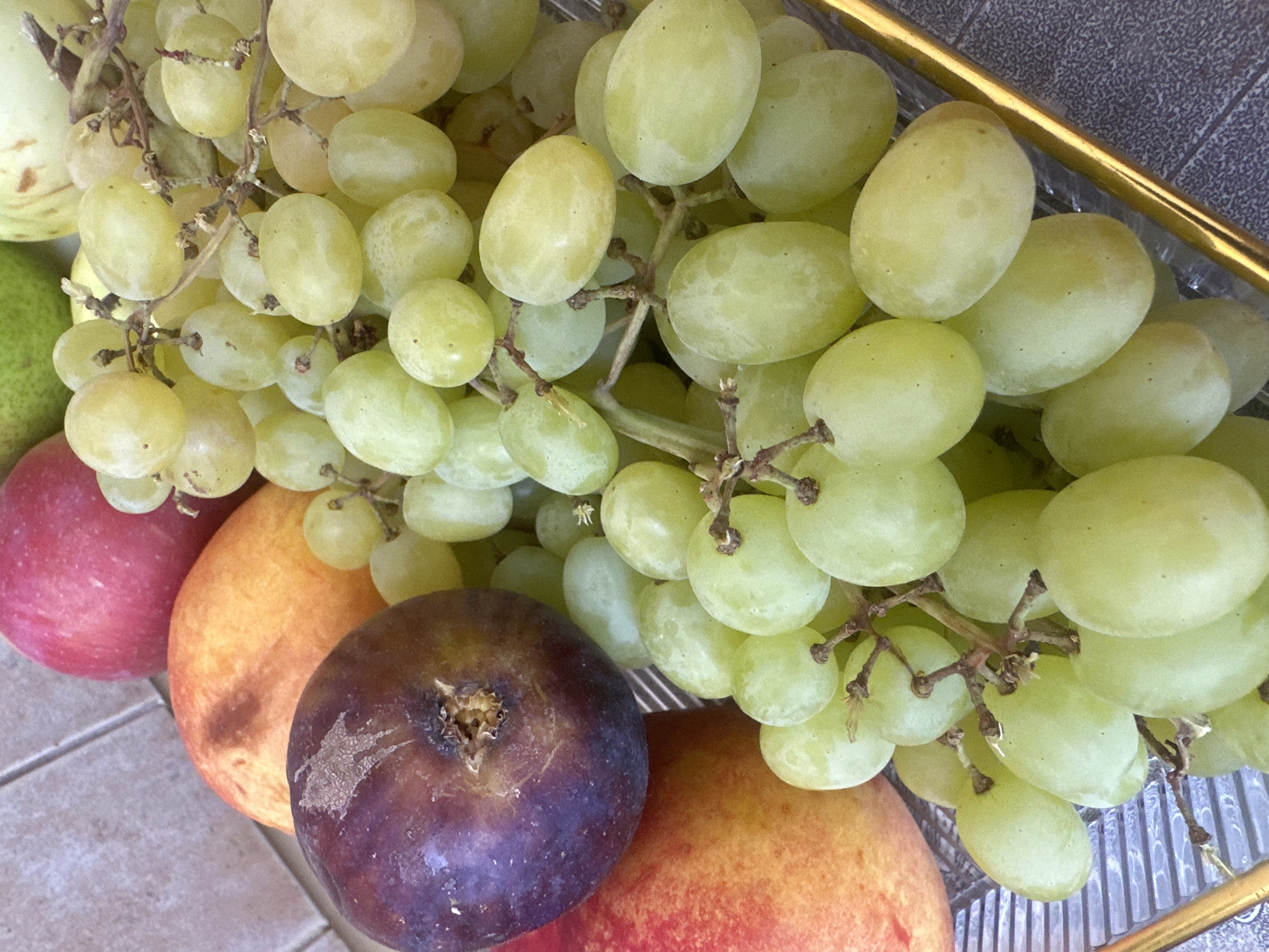 A close-up, overhead shot displays a collection of fresh fruits, primarily green grapes, artfully arranged on a transparent tray with a gilded edge. Interspersed among the grapes are a deep purple fig, a ripe peach with a gradient of yellow to red, and a bright red apple. The fruits are partially resting on a tiled surface. The image does not contain any information that would allow for the identification of its location within the city of Ternopil, Ukraine.