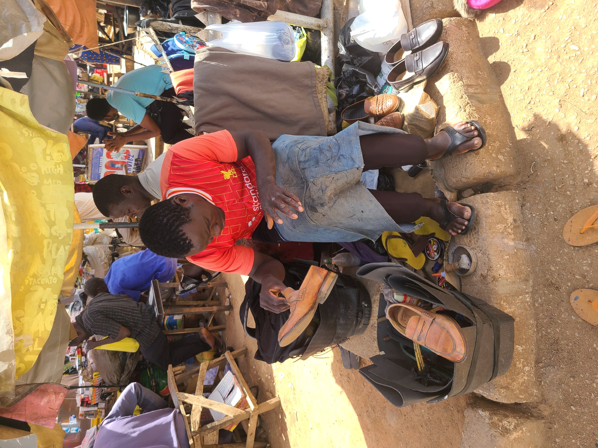 The scene depicts a shoe repair service in an outdoor, bustling market setting in the city of Unknown, Unknown. A man wearing a red shirt with the Liverpool FC logo and denim shorts is holding a brown leather loafer in his right hand, presumably working on it. He is sitting amidst his tools and a black bag containing more shoes and shoe-related supplies. Next to him, on what appears to be a concrete structure, are several pairs of shoes and sandals. In the background, there are other individuals, some seated and seemingly engaged in similar or related activities under makeshift shelters. The environment is crowded, with an assortment of goods and materials visible, suggesting a vibrant and active marketplace.