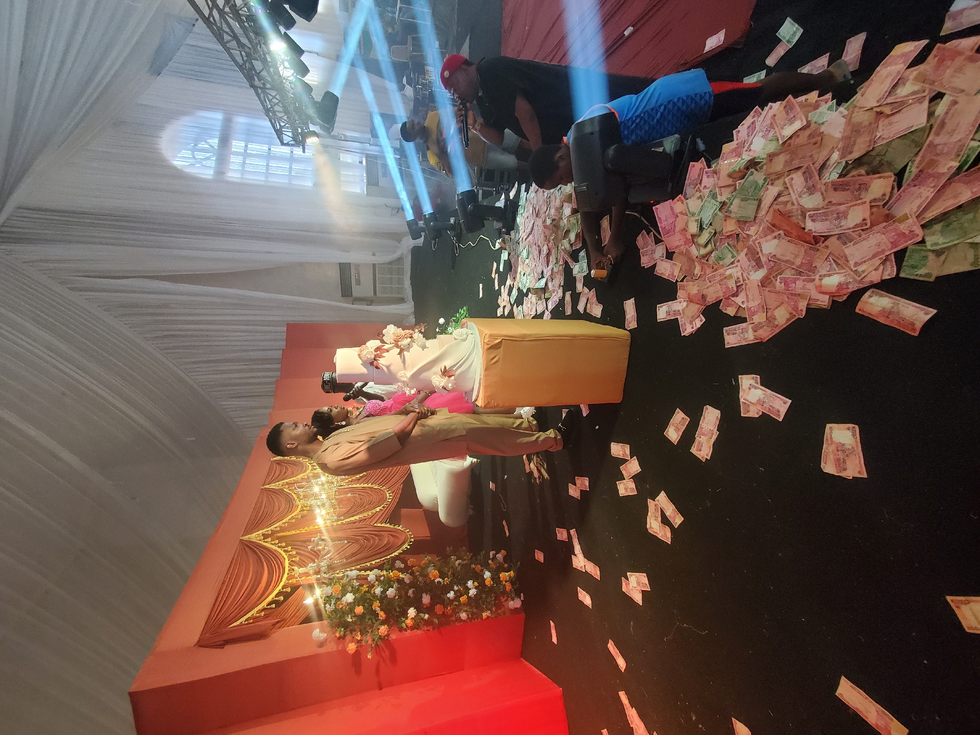 The image depicts a scene likely at a wedding reception or a similar celebratory event. The location is Unknown, Unknown. A couple stands on a decorated stage with an orange and white motif. A pile of banknotes, predominantly red and white, is scattered on the floor. A group of people including a man in a red hat, and a man in a blue shirt, are picking up the banknotes. Lighting equipment is visible in the ceiling of the venue.
