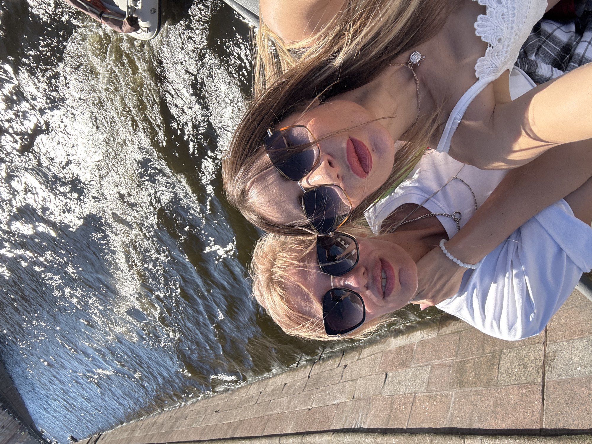Two women are taking a selfie on a boat on a sunny day. The woman in the foreground on the right has long blonde hair, is wearing sunglasses, and has pink lipstick on. The woman behind her on the left has blonde hair and is wearing sunglasses and a white t-shirt. The boat is on a body of water, likely a canal or river, with a stone embankment on the left side. The water is choppy and reflecting the sunlight. The overall scene suggests a leisurely outing in Saint Petersburg, Russia, possibly along the Neva River or one of its many canals, given the architectural style of the embankment.