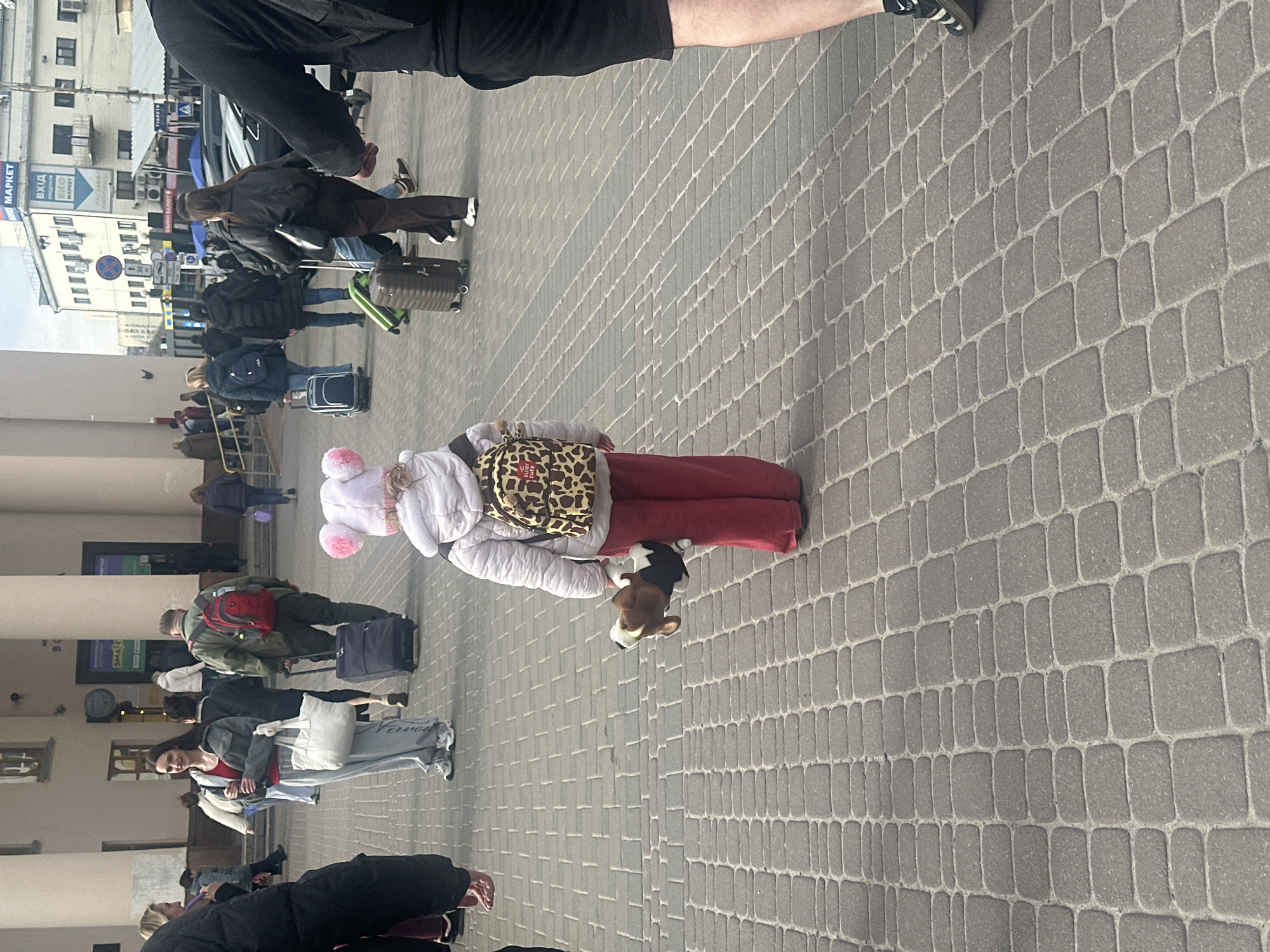 The image shows a bustling outdoor scene in Kyiv, Ukraine, on a paved pedestrian area. Numerous people are present, appearing to be in transit or waiting, with many carrying luggage. The lighting suggests it is daytime, possibly overcast.
In the foreground, a child is prominently featured, dressed in a white puffer jacket, red pants, and a white hat adorned with two large pink pom-poms. The child wears a leopard-print backpack, which has a small red tag with the Ukrainian word "КРИХІТКА" (KRYKHITKA, meaning "Baby" or "Tiny Tot"). The child is holding a plush black, white, and brown dog toy.
Behind the child, several adults are visible. Many are equipped with various types of luggage, including roller suitcases, backpacks, and carry-on bags. One woman in the mid-ground wears a grey sweatshirt and light grey sweatpants, carrying a white tote bag with the text "Vorova" visible on its side. Other individuals further back are also seen with luggage, suggesting travel.
The ground is covered with dark grey, rectangular interlocking paving stones. In the background, multi-story buildings line the street. A blue sign on one of the buildings is partially visible, showing the word "MARKET" and the Ukrainian word "ВХІД" (VKHID), meaning "Entrance." Other street signs, largely unreadable, are also present. The scene conveys a sense of movement and waiting, likely at a transportation hub.
