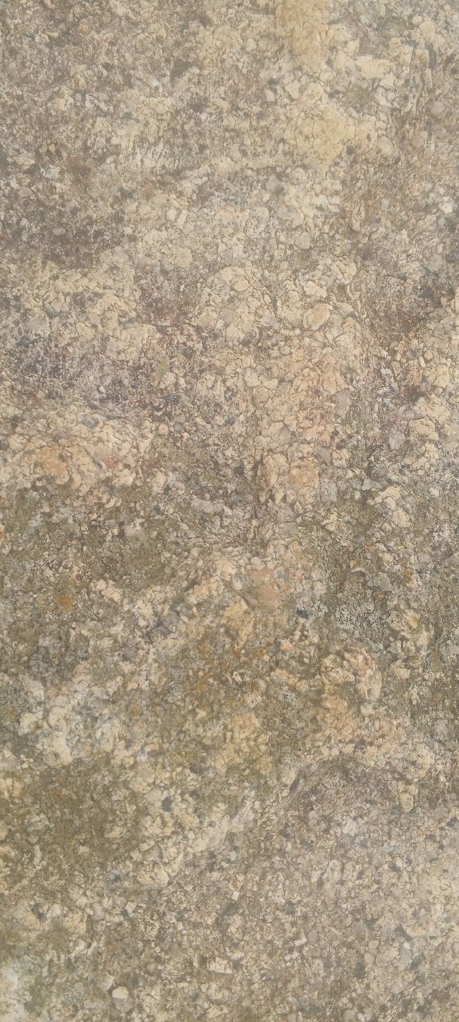 This is a close-up, top-down view of a textured surface that appears to be a natural stone or concrete with a speckled, uneven pattern. The colors range from light beige and cream to various shades of brown and gray. The texture is rough and granular, with small, irregular fragments visible. The lighting suggests an outdoor environment, likely in Ohit, Nigeria, given the context. There are no people or specific objects clearly identifiable in the image, only the textured surface itself. The overall impression is of a weathered, earthy material.