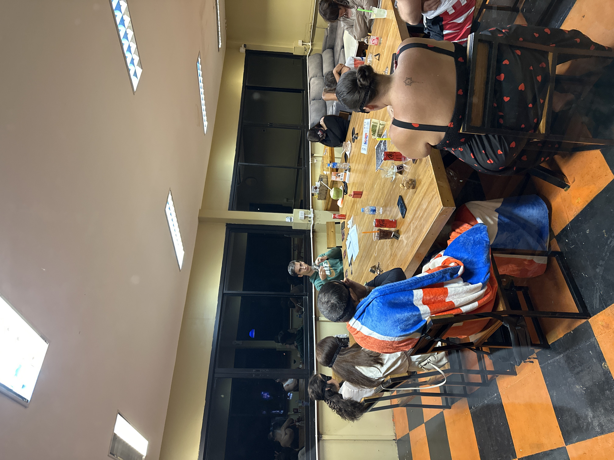 In an indoor room with fluorescent lighting and yellow walls, a group of ten people are gathered around a large wooden table, some with black eye masks on. A man in a green hoodie sits at the head of the table, facing away from the camera. The room has a checkered orange and black floor and large windows looking out onto a dark exterior. On the table are various items including cups, bottles, a coconut, and what appear to be playing cards or game pieces. The people are engaged in an activity around the table, with some looking at the table or each other, and others with their heads down.
